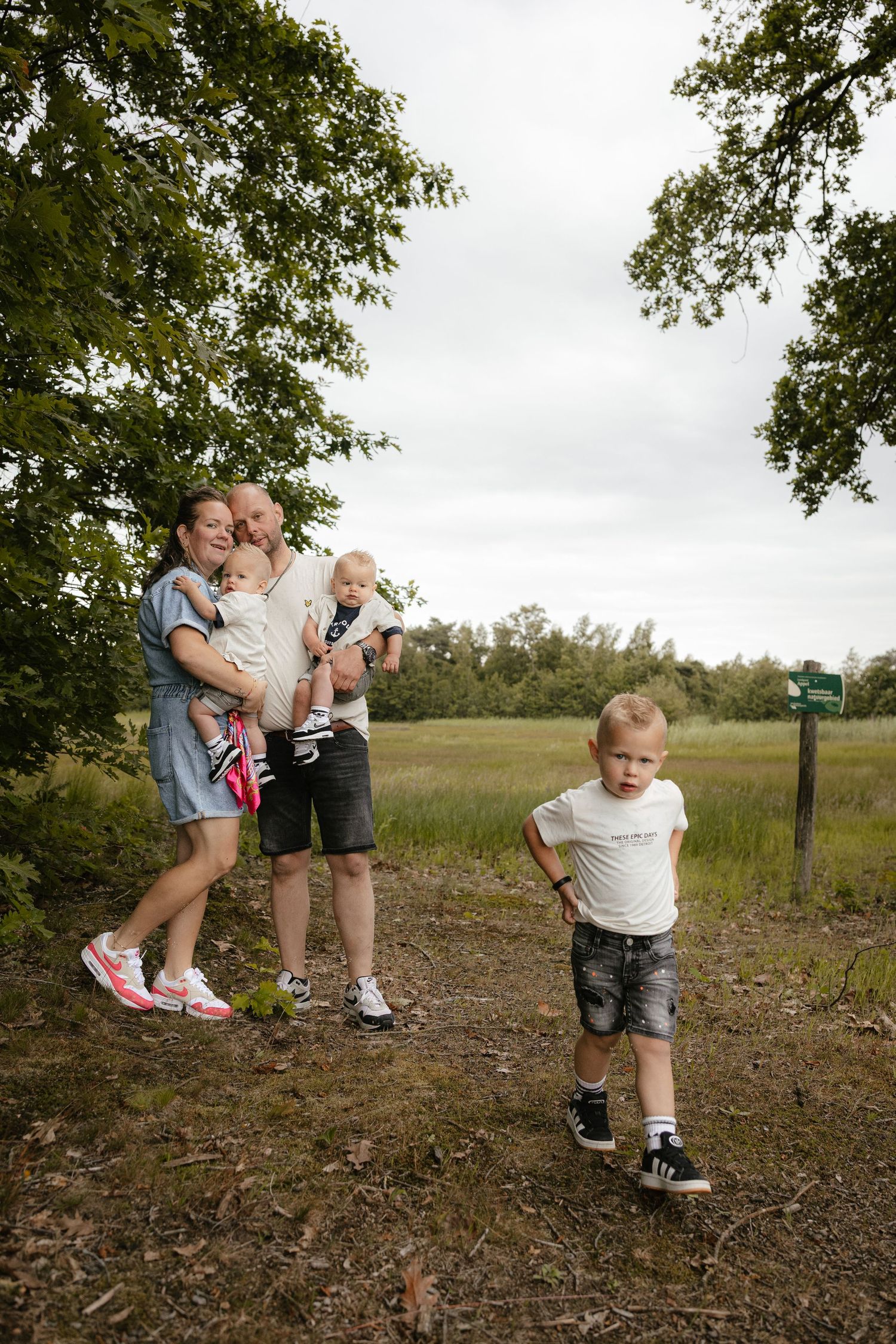 Family with young children posing outdoors on a nature trail, with trees and green meadow in the background.