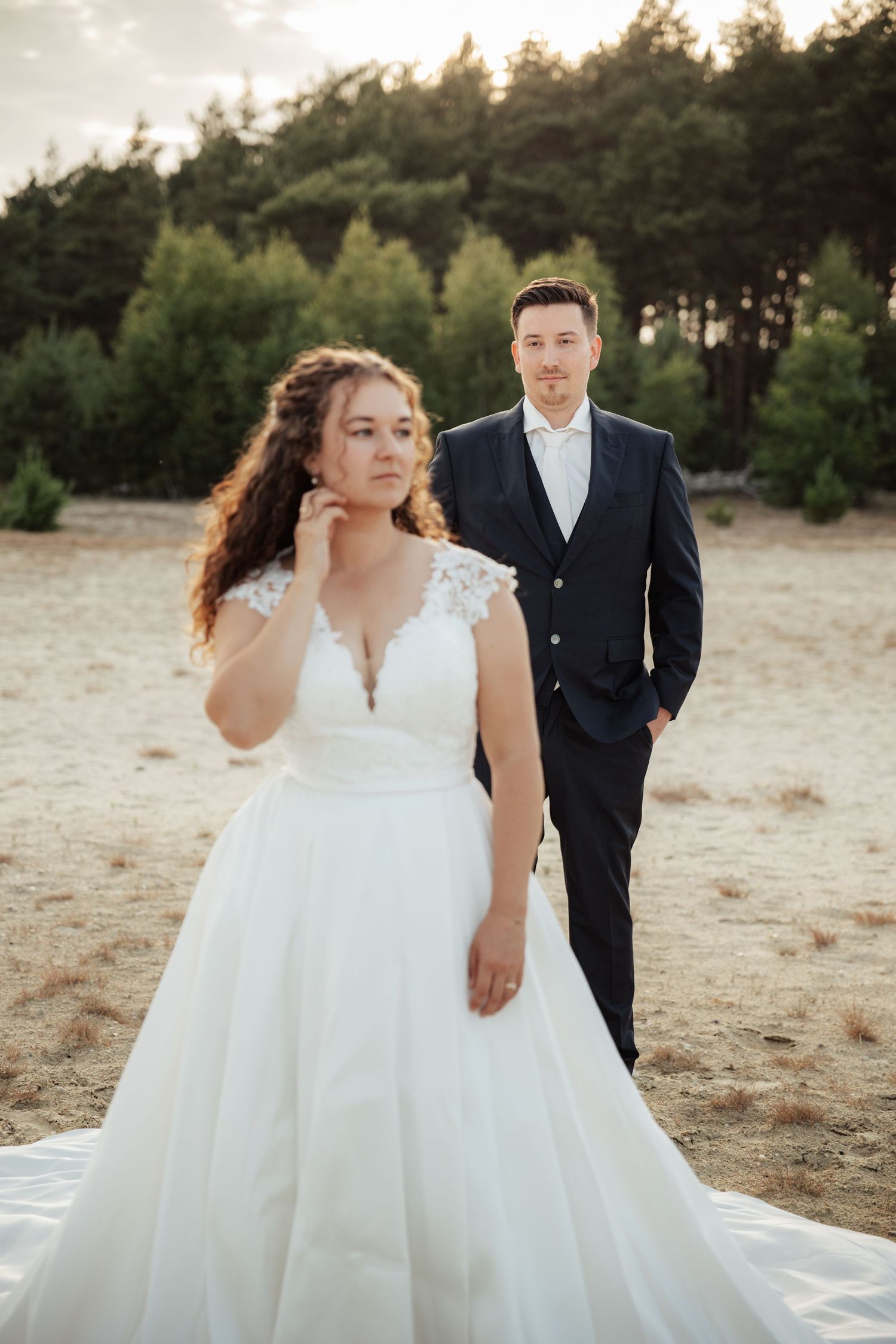 Bride in white lace gown and groom in navy suit pose outdoors on sandy ground with pine forest backdrop at sunset.