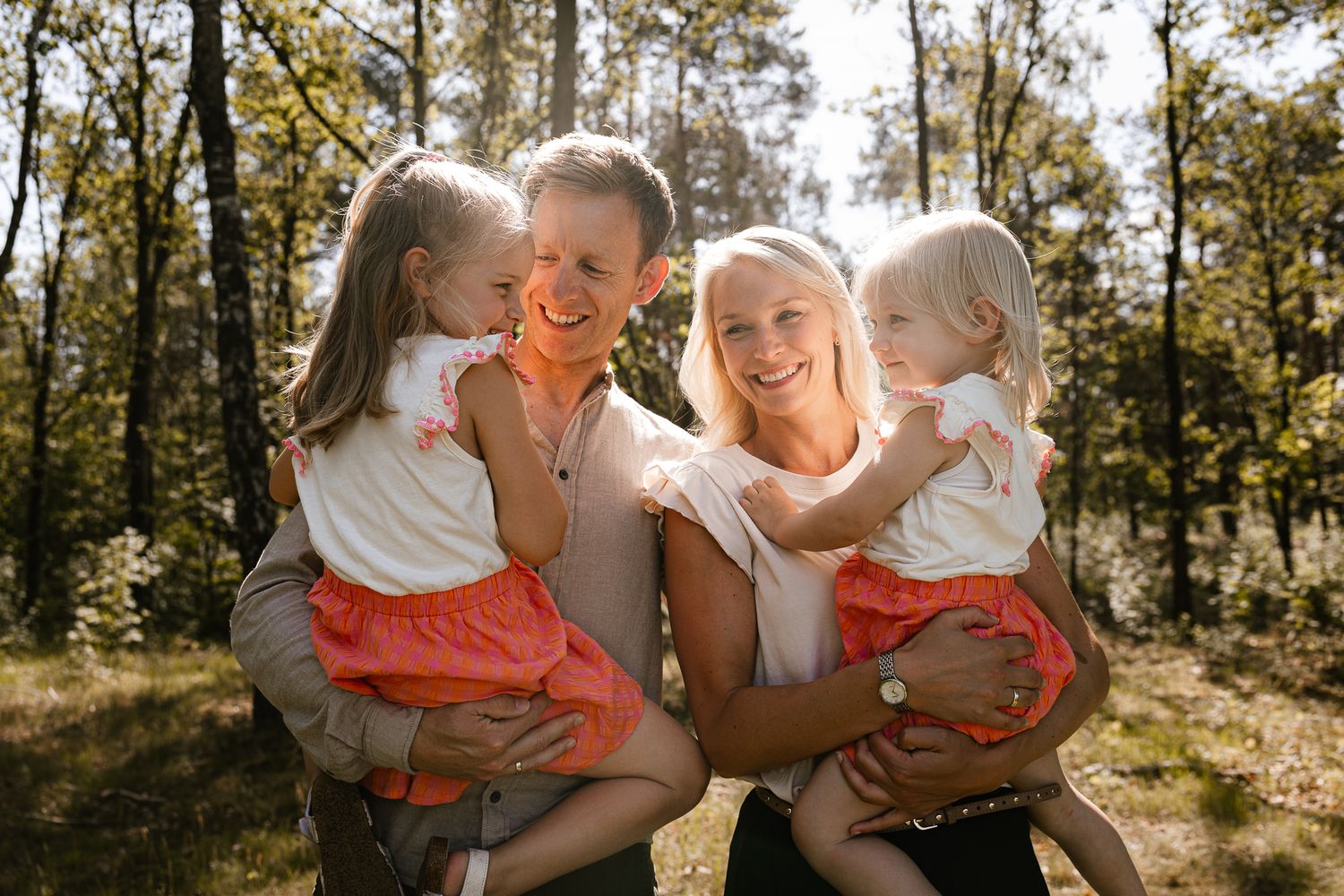 Happy family of four posing outdoors in a sunny forest, parents holding two young girls in matching red skirts.