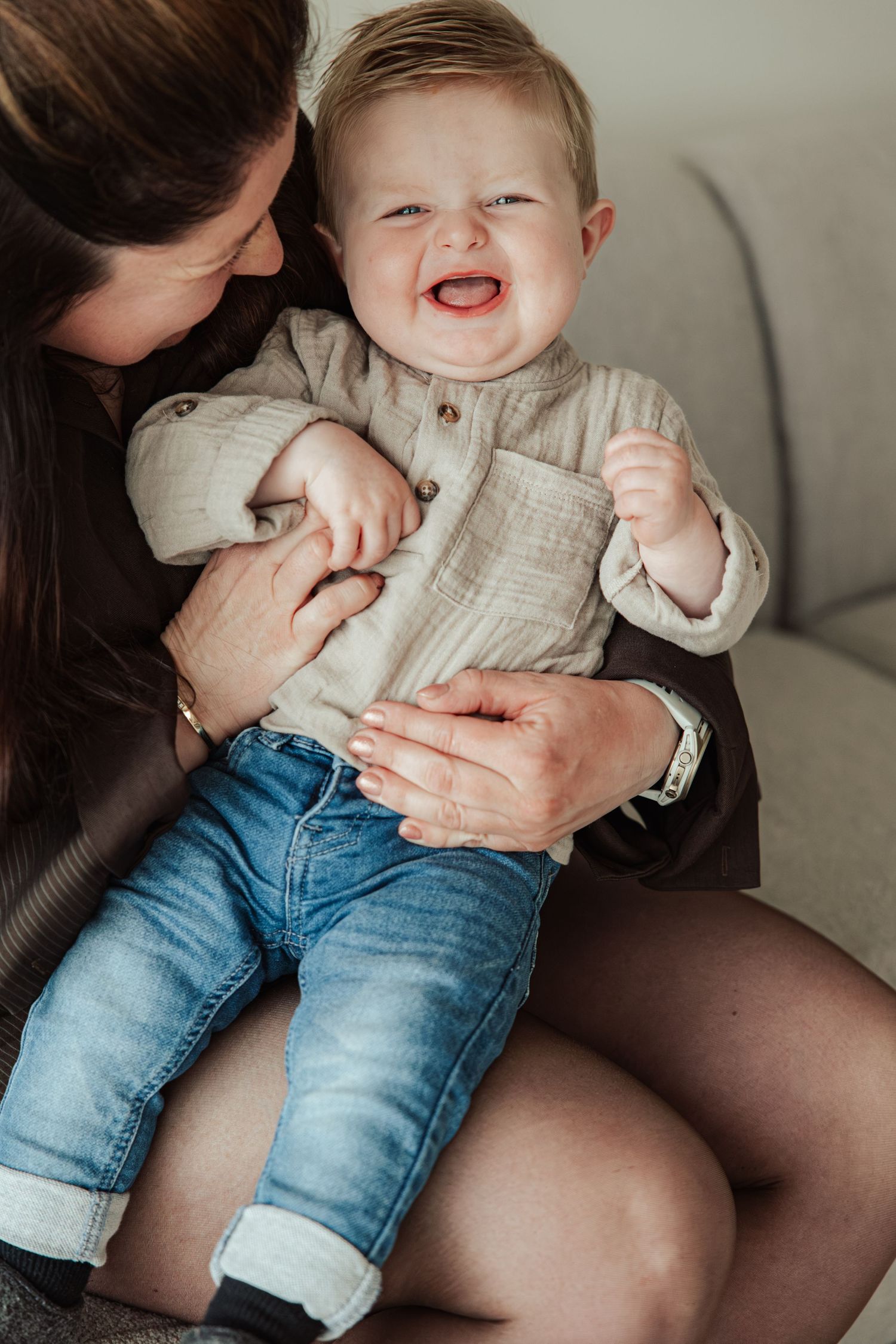 Happy baby boy laughing while being held by his mother on a couch, wearing jeans and a beige linen shirt.