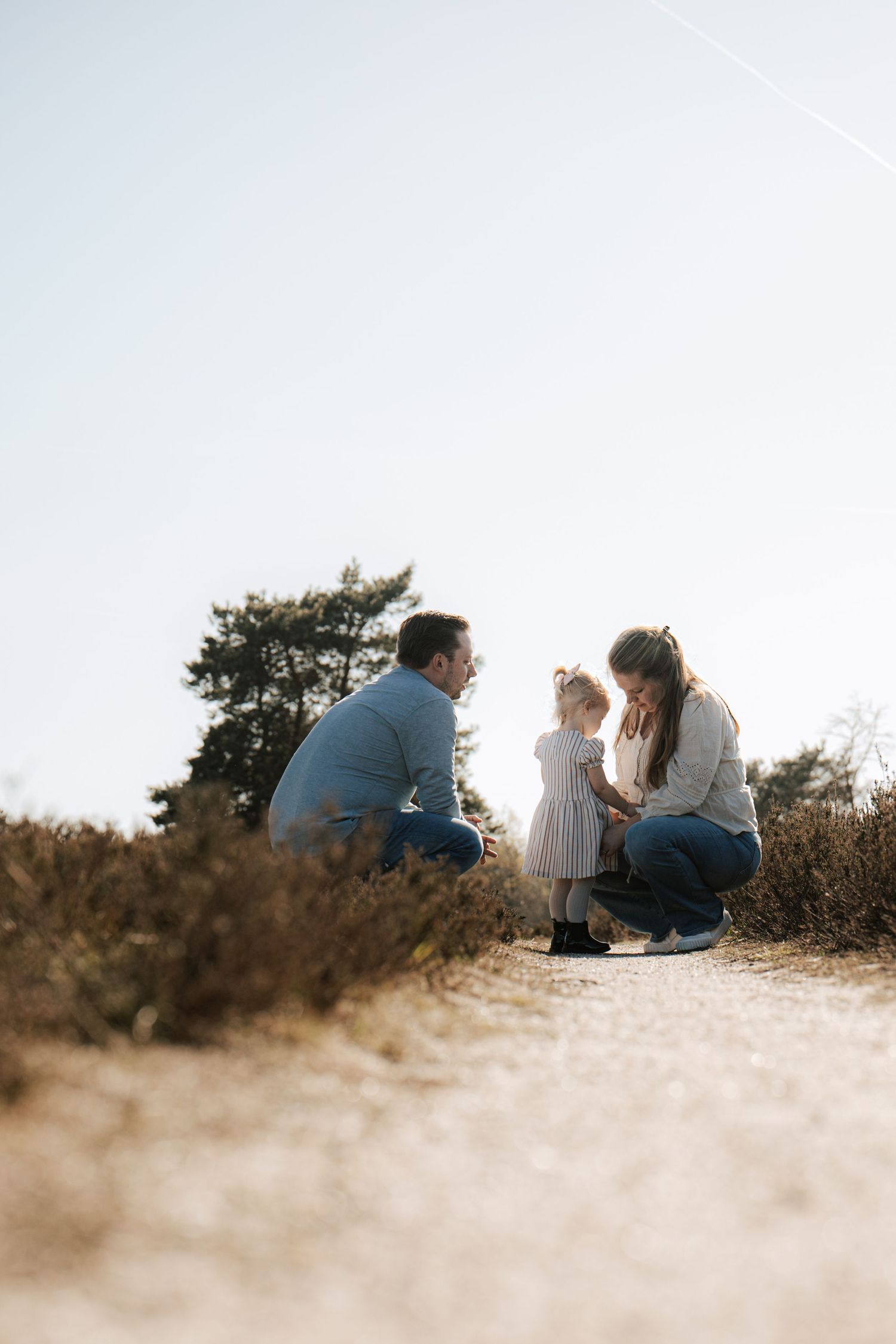 A family with a young toddler crouching together on a sunny dirt path surrounded by golden heathland.