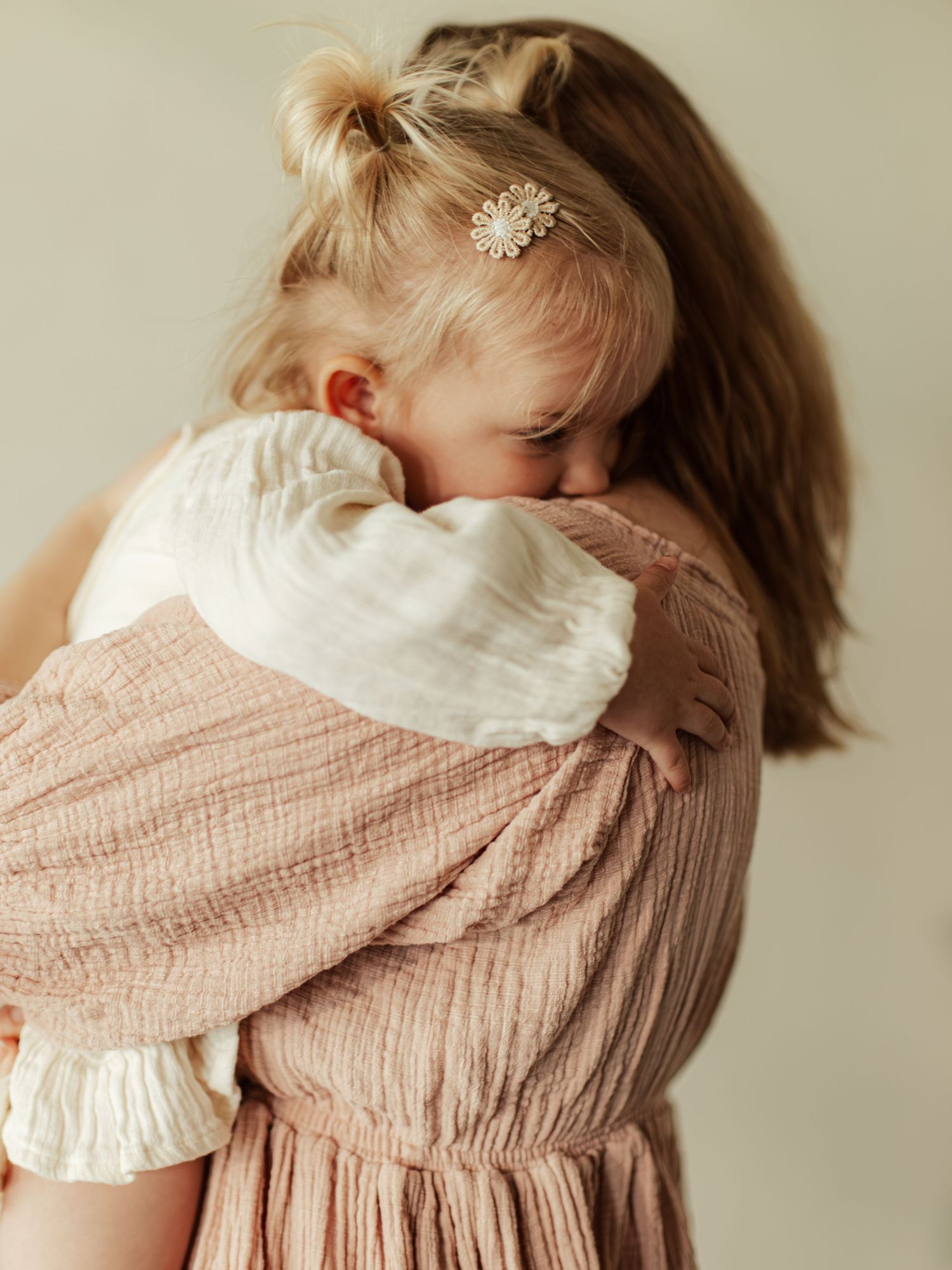 A young toddler girl with a floral hair clip hugs a woman in a pink dress in a tender embrace.