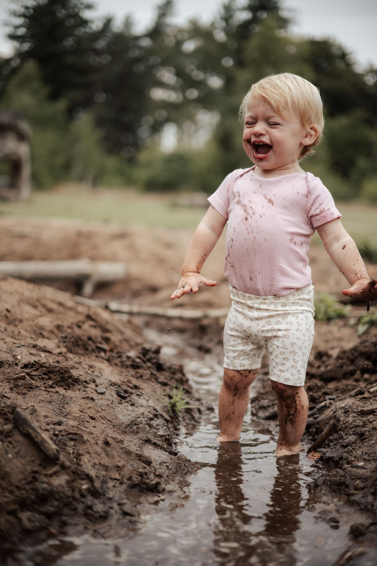 Toddler in pink shirt and patterned shorts splashing joyfully in muddy puddles.
