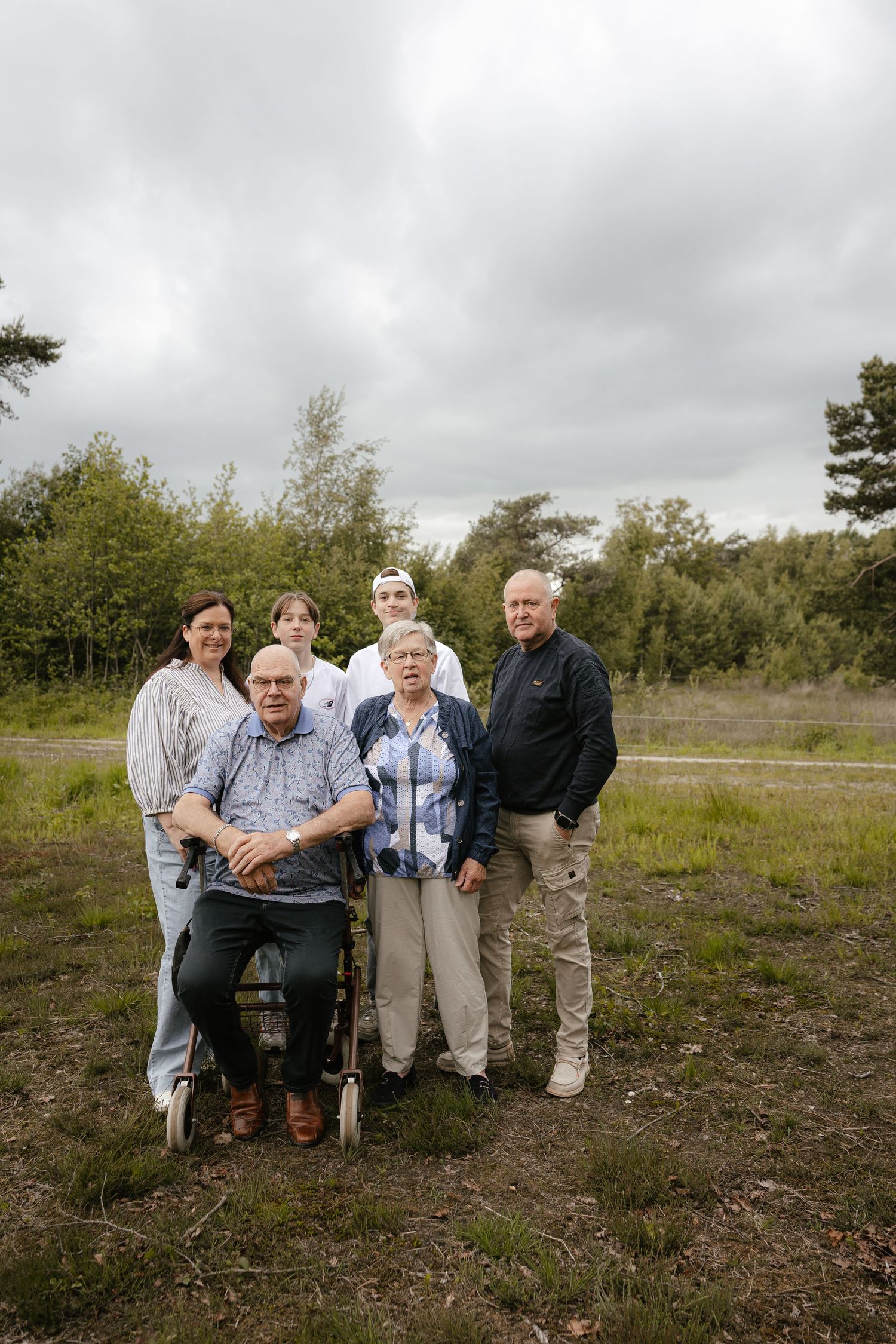 Family group portrait outdoors in nature, including elderly man in wheelchair, gathered on grassy field with trees and cloudy sky.