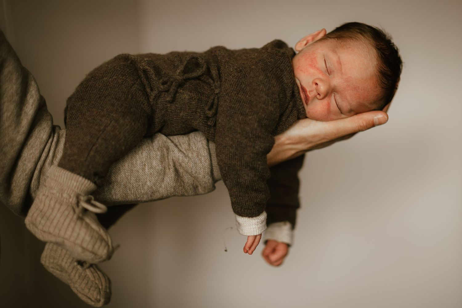 Sleeping newborn baby in brown knit outfit cradled in adult hands against neutral background.