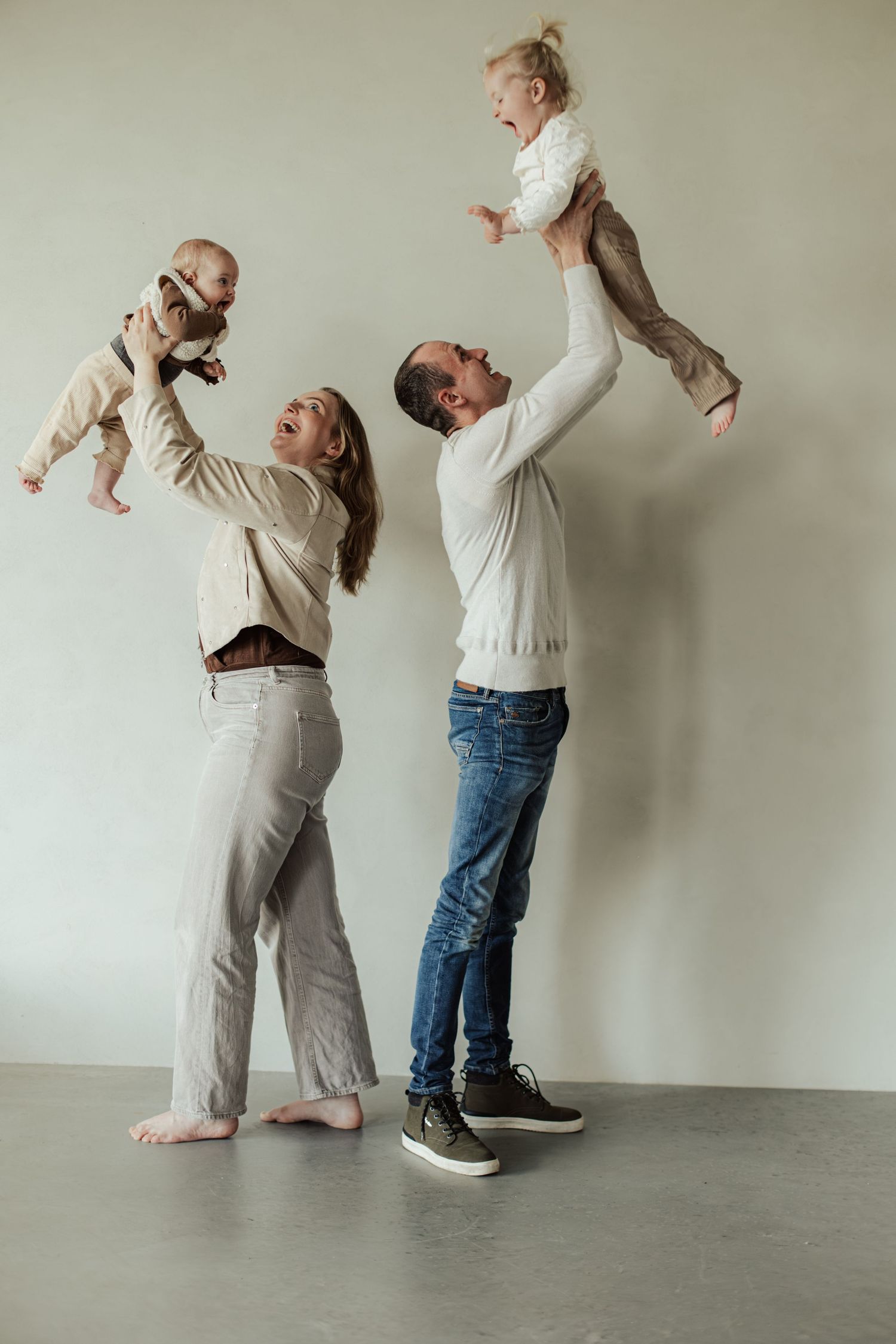 Happy family with two parents joyfully lifting their babies in the air against a light gray wall.
