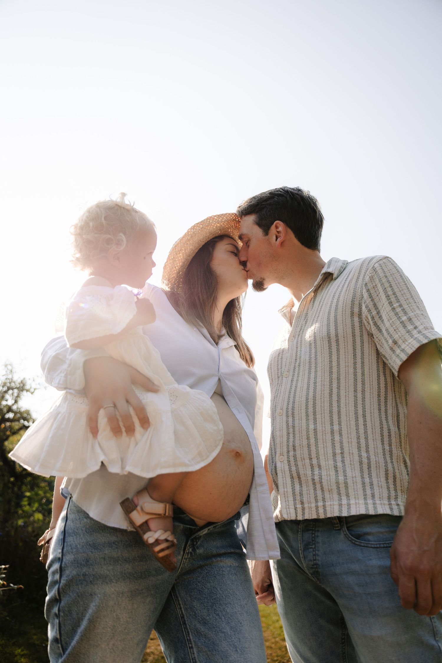 Pregnant woman and man kissing outdoors in sunlight while holding a toddler girl in a white dress.