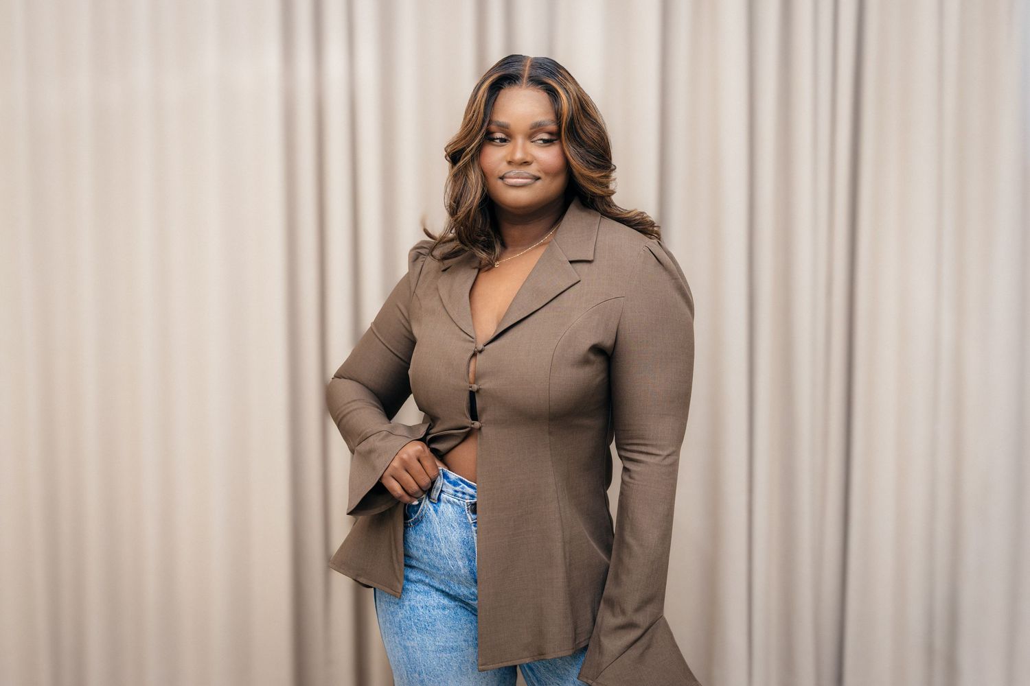 Fashion model poses confidently in brown button-up top and jeans against neutral curtain background.