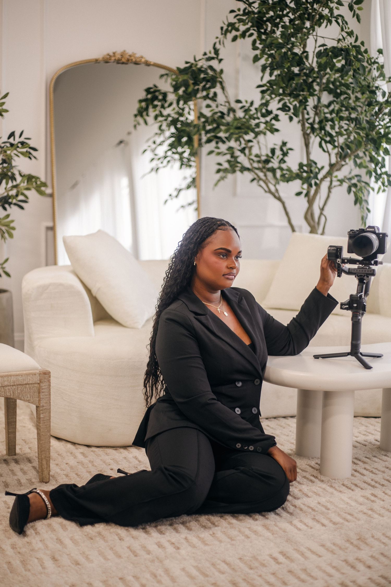 A woman in a black suit sits on a floor, adjusting a camera on a gimbal tripod in a bright, modern living room.