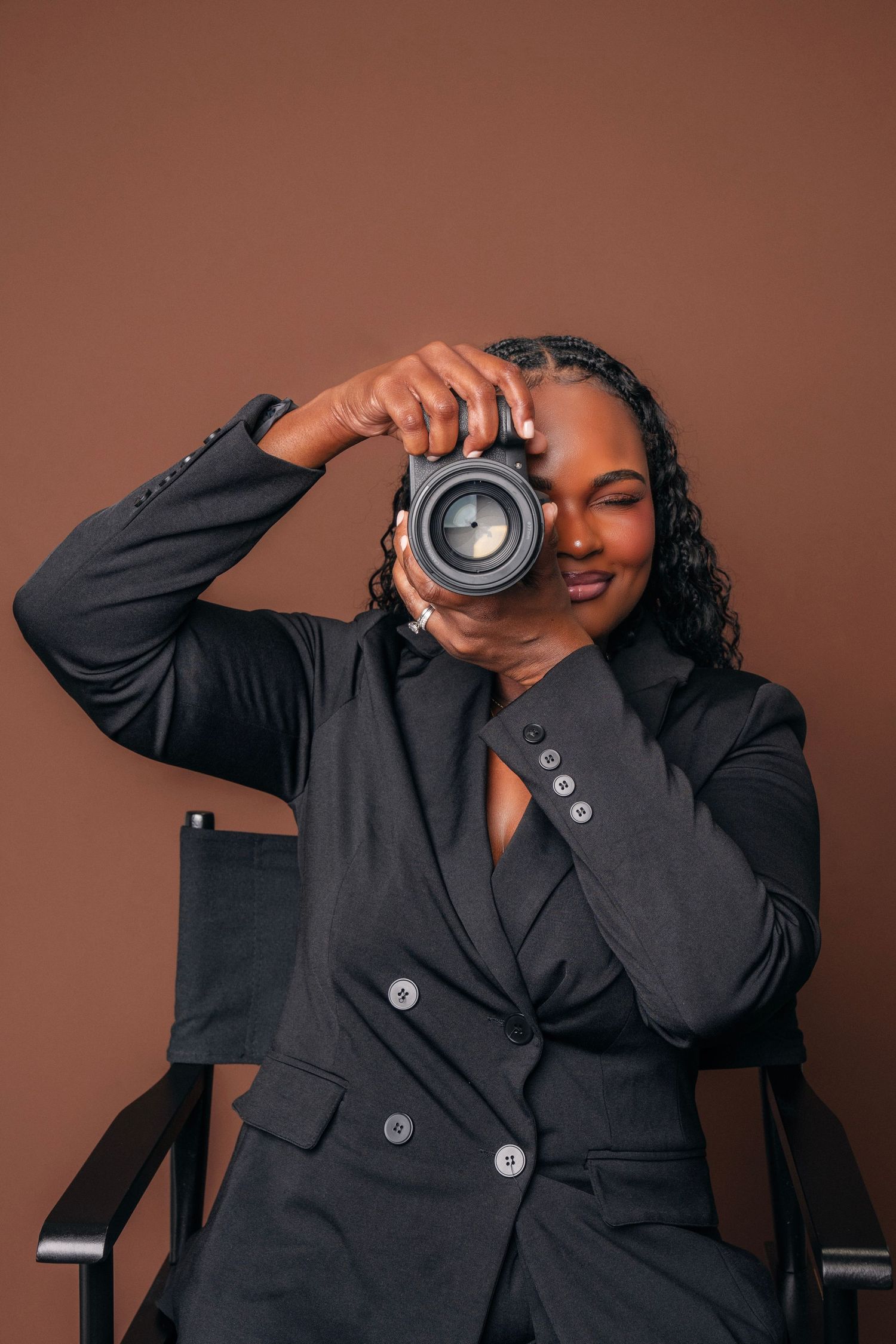 A professional Black woman photographer in a black blazer sits in a director's chair, holding a camera up to her eye.