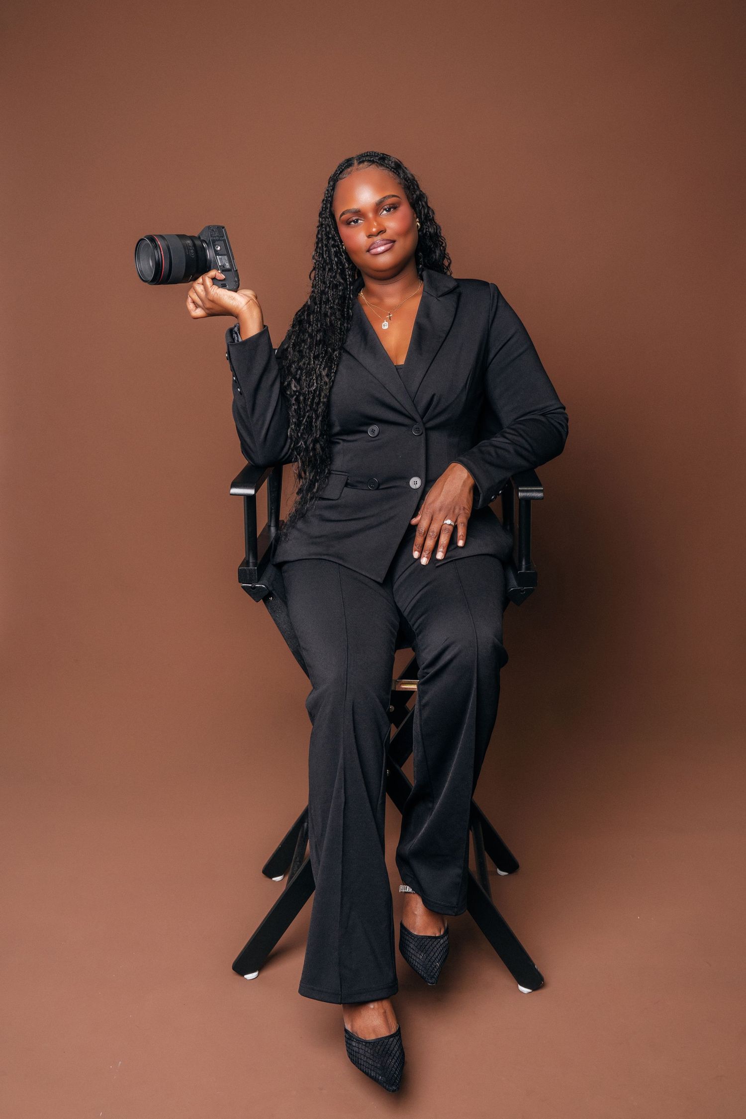 A professional Black woman photographer in a black suit sits in a director's chair holding a DSLR camera against a brown backdrop.