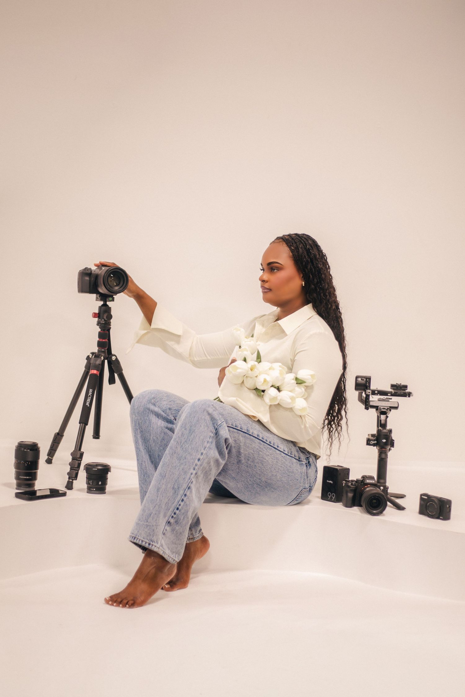 A woman in white shirt and jeans sits barefoot holding white flowers, surrounded by cameras and tripods in a studio.
