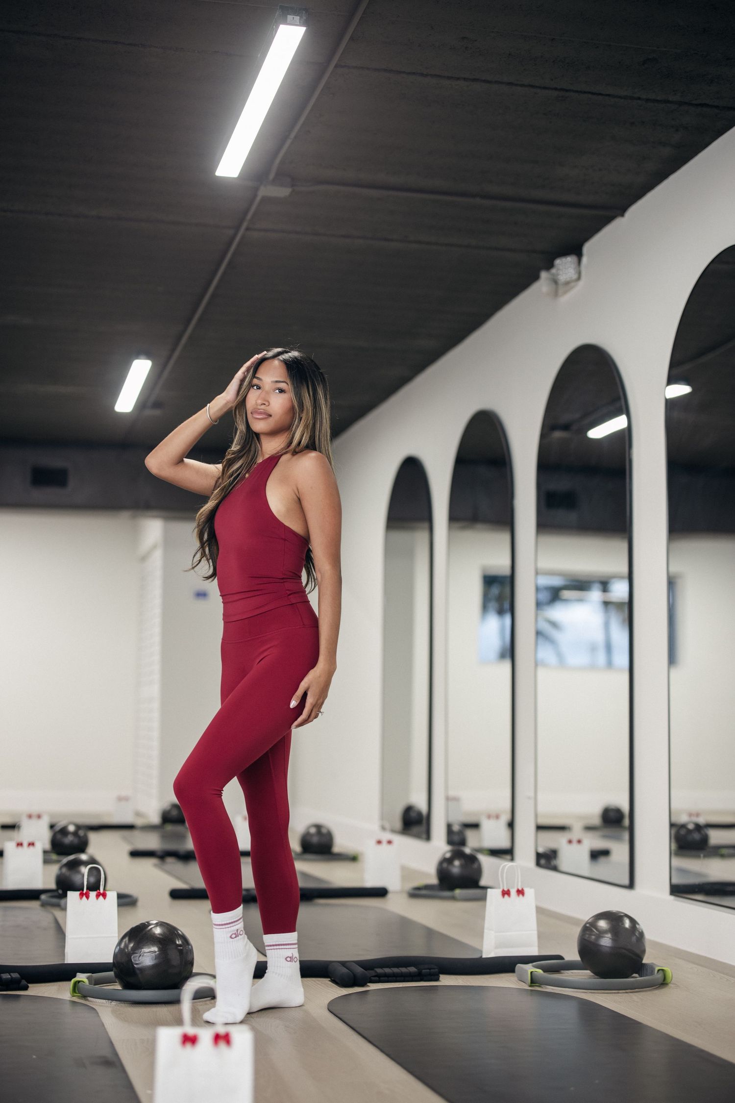 A person in burgundy athletic wear performs stretching exercises in a modern gym space with fluorescent lighting.