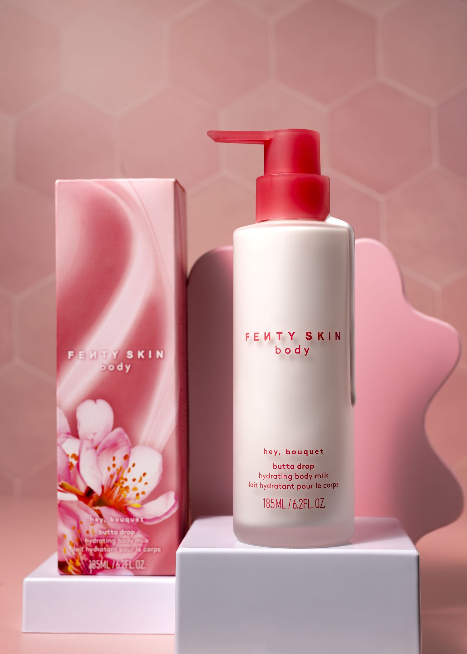 Fenty Skin Body Hey Bouquet Butta Drop hydrating body milk pump bottle beside pink floral box on white pedestal.