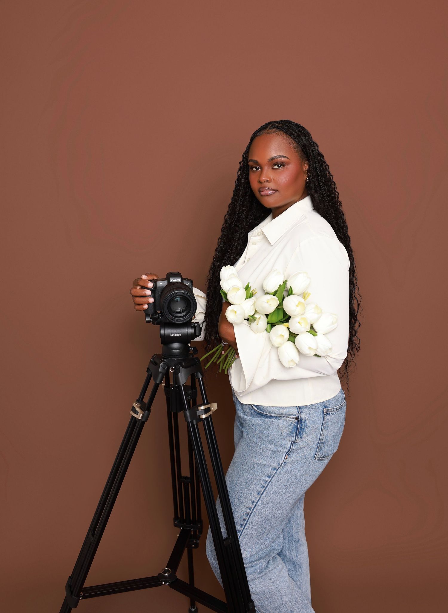 A woman photographer poses with a camera on a tripod, holding white tulips, wearing a white jacket and jeans.