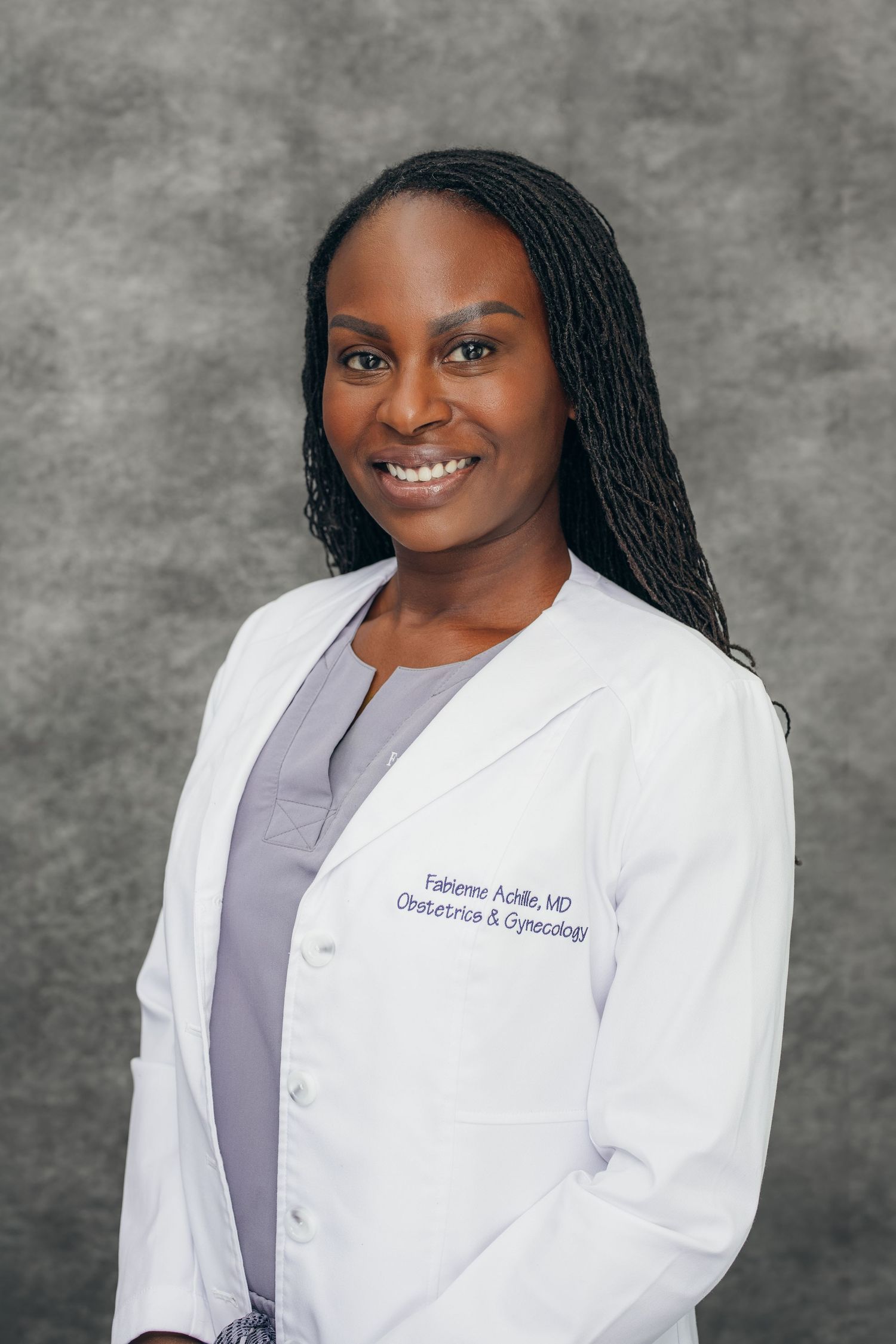 Female OB-GYN physician Fabienne Achille, MD, smiling in a white lab coat against a gray background.