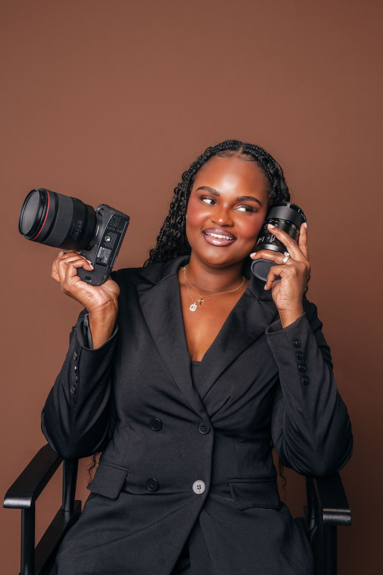 Smiling Black female photographer in black blazer holding DSLR cameras while seated against a brown studio background.