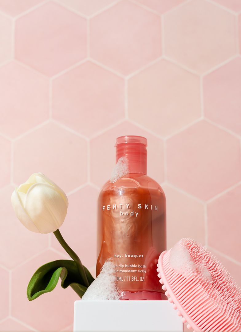 Fenty Skin Body rose bouquet bubble bath bottle displayed with a white tulip and pink scrubber on a white pedestal.