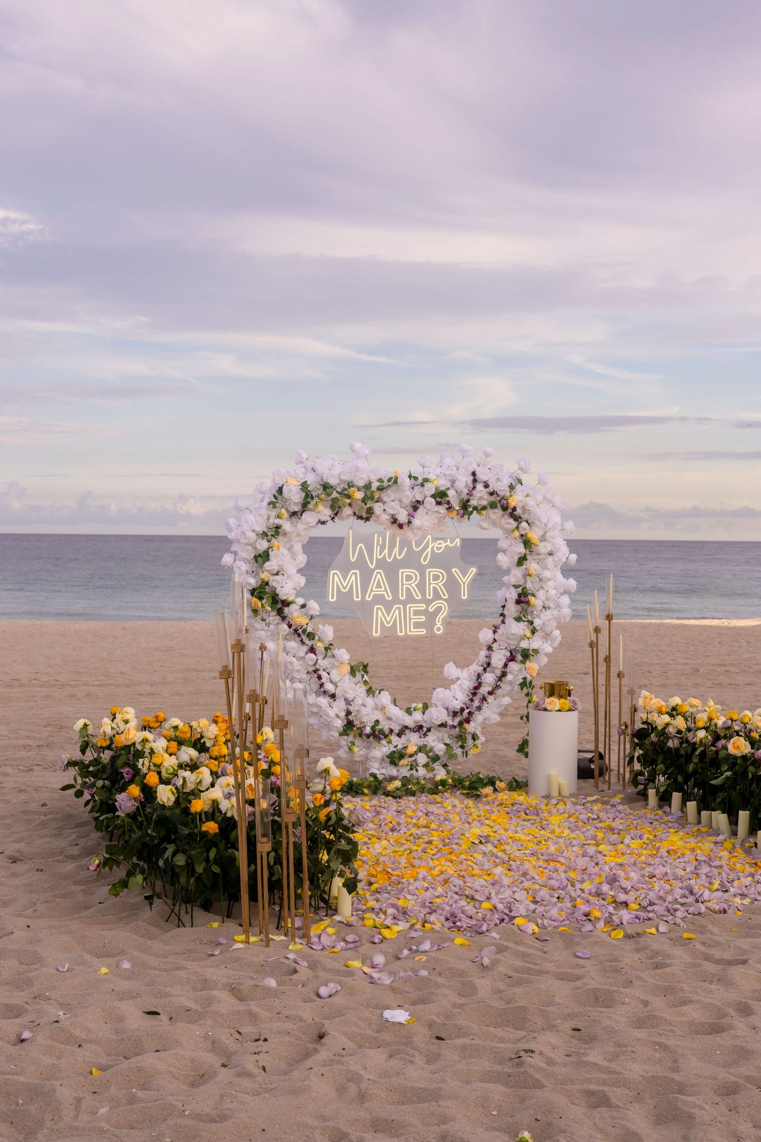 Beach proposal setup with illuminated sign, floral arch, and yellow petals on sand at sunset.