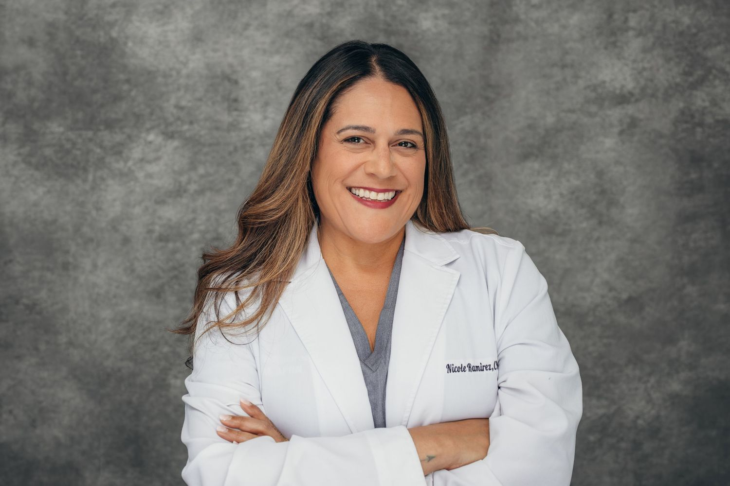 Smiling female doctor in white lab coat with arms crossed poses professionally against gray studio background.