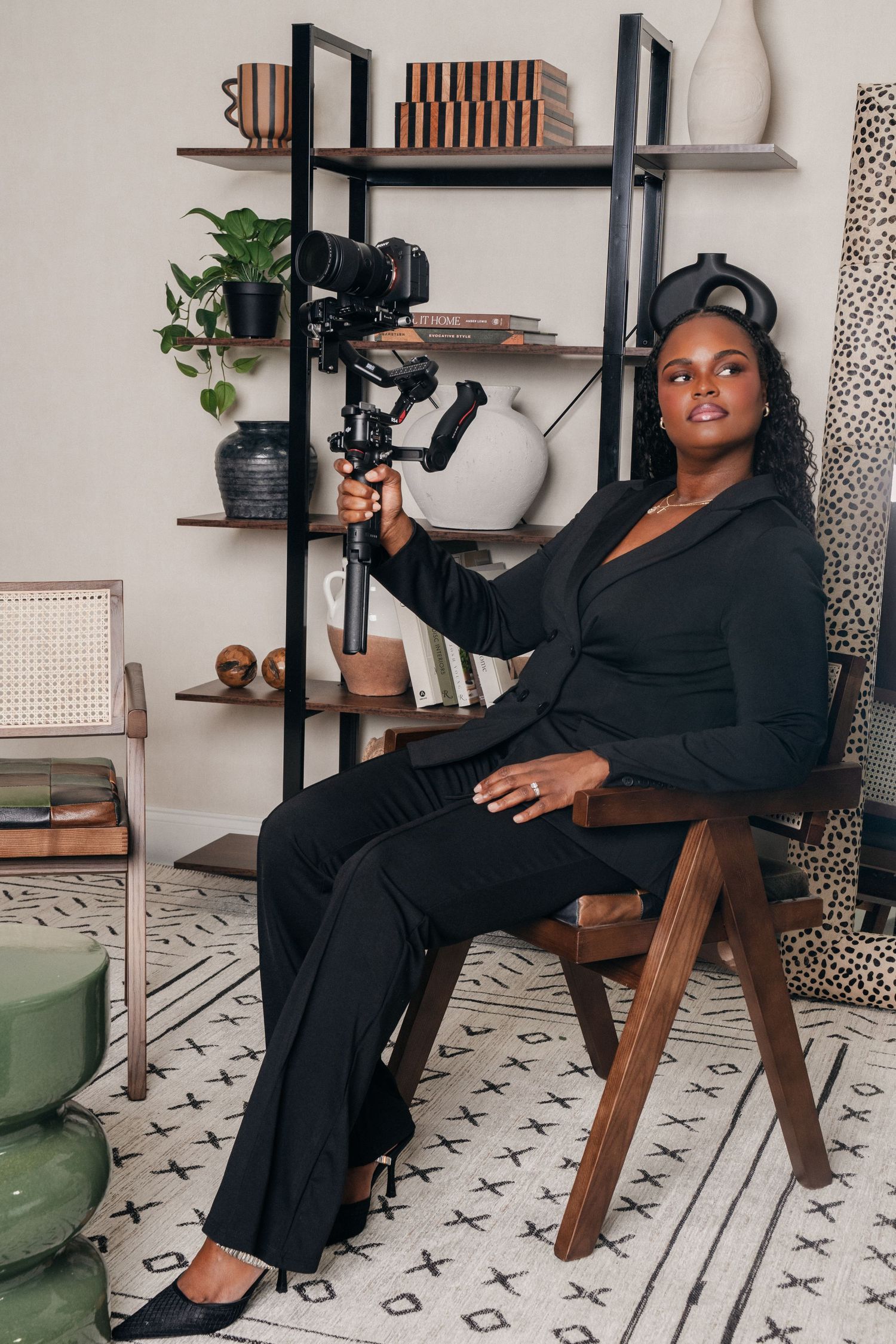 A woman in a black suit sits in a wooden chair holding a camera gimbal stabilizer in a stylish modern home setting.