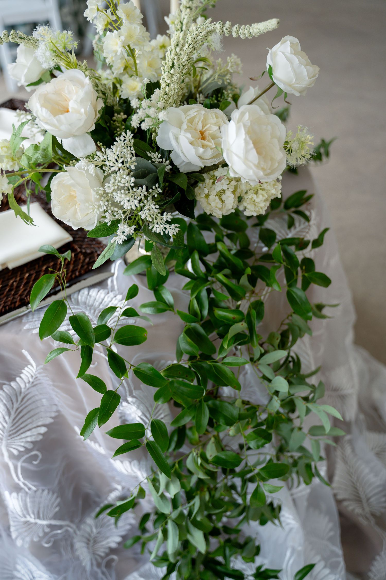 Elegant wedding table with white roses, greenery garland, and floral arrangements on a white embroidered tablecloth.
