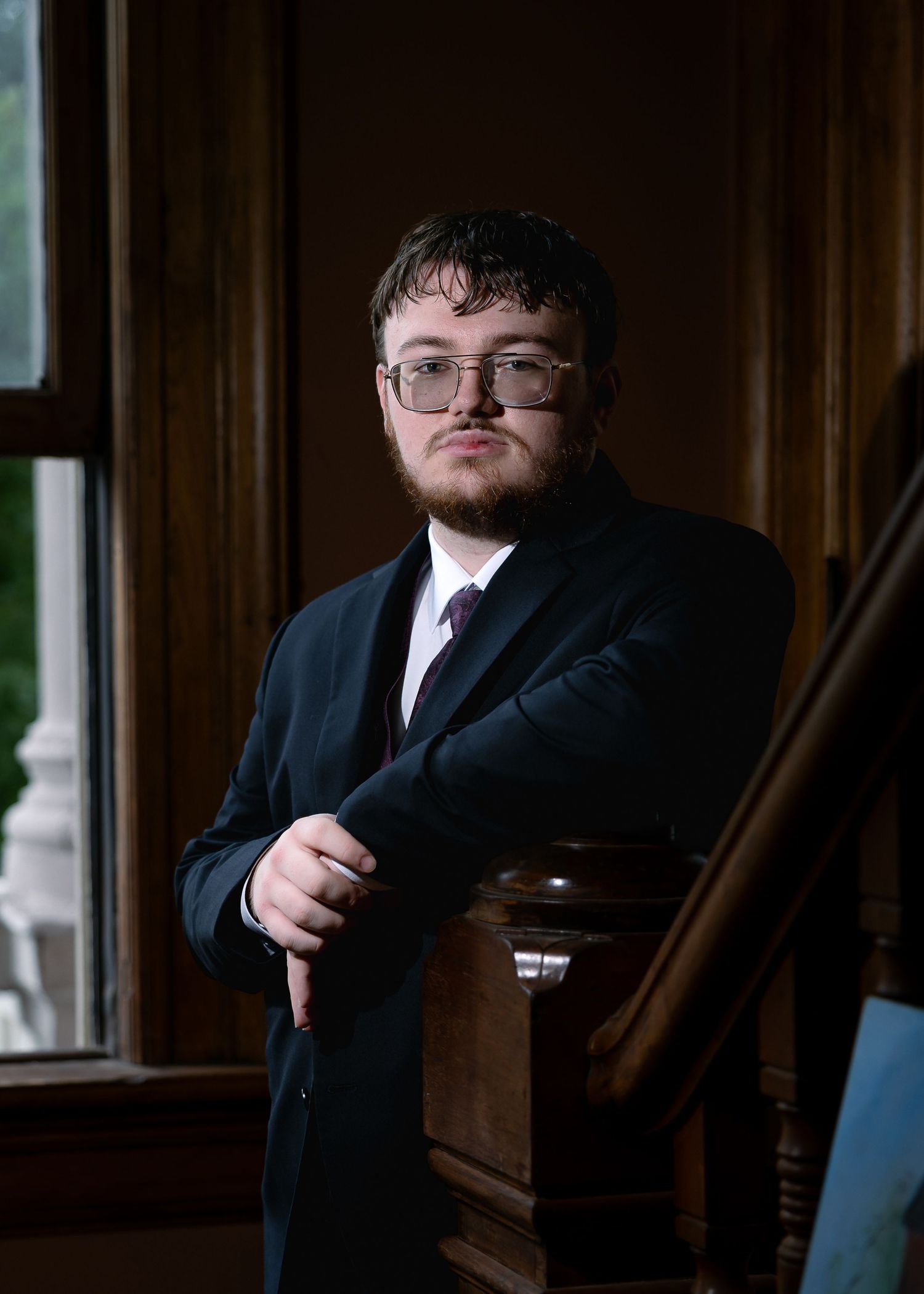 Young man in black suit and glasses leaning on wooden staircase railing in elegant interior setting.