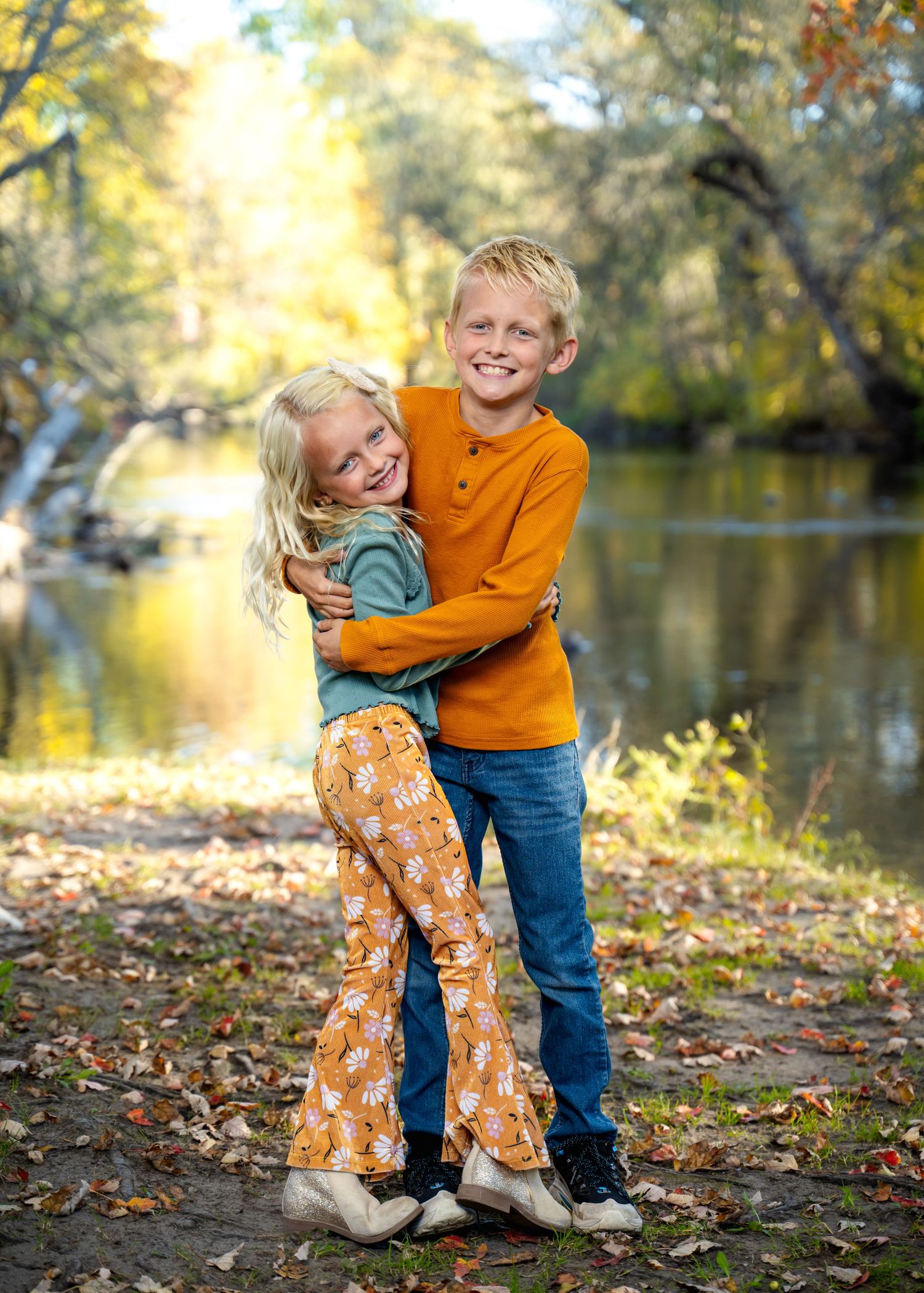 Two children share a warm hug while standing near a lake surrounded by fall foliage.