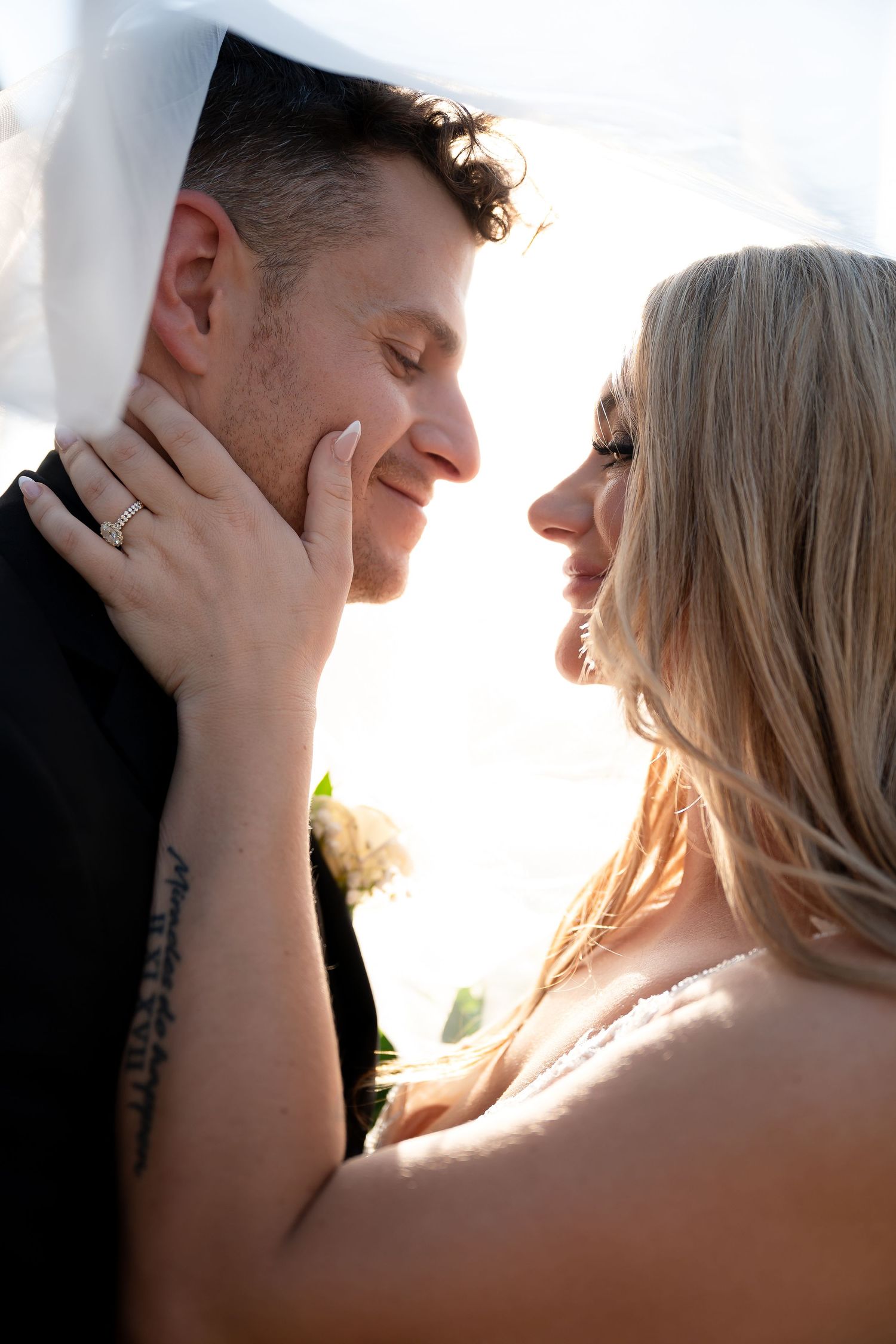 A smiling bride and groom share an intimate moment under a veil, with the bride's diamond ring visible on her hand.