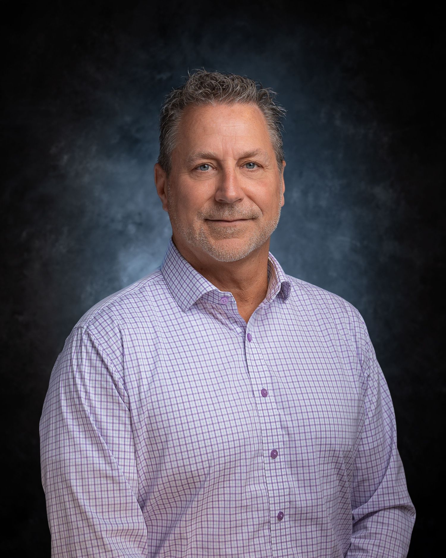 Professional headshot of a smiling middle-aged man in a light purple checkered shirt against a dark studio background.