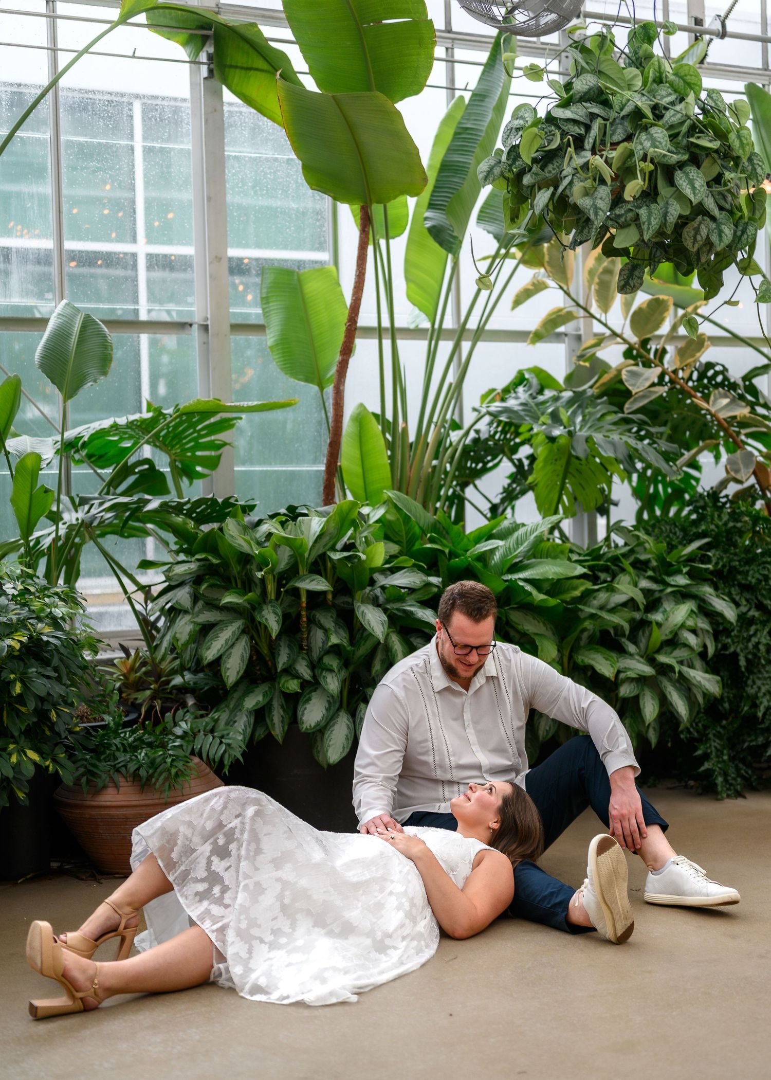 Two people lying on floor in indoor garden setting with lush green plants and natural lighting.