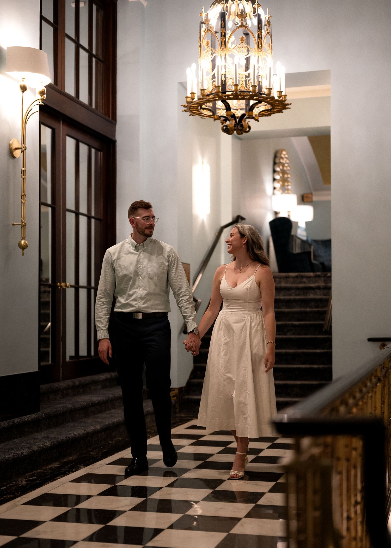 Couple holding hands walking through elegant hotel lobby with chandelier and black and white checkered floors.