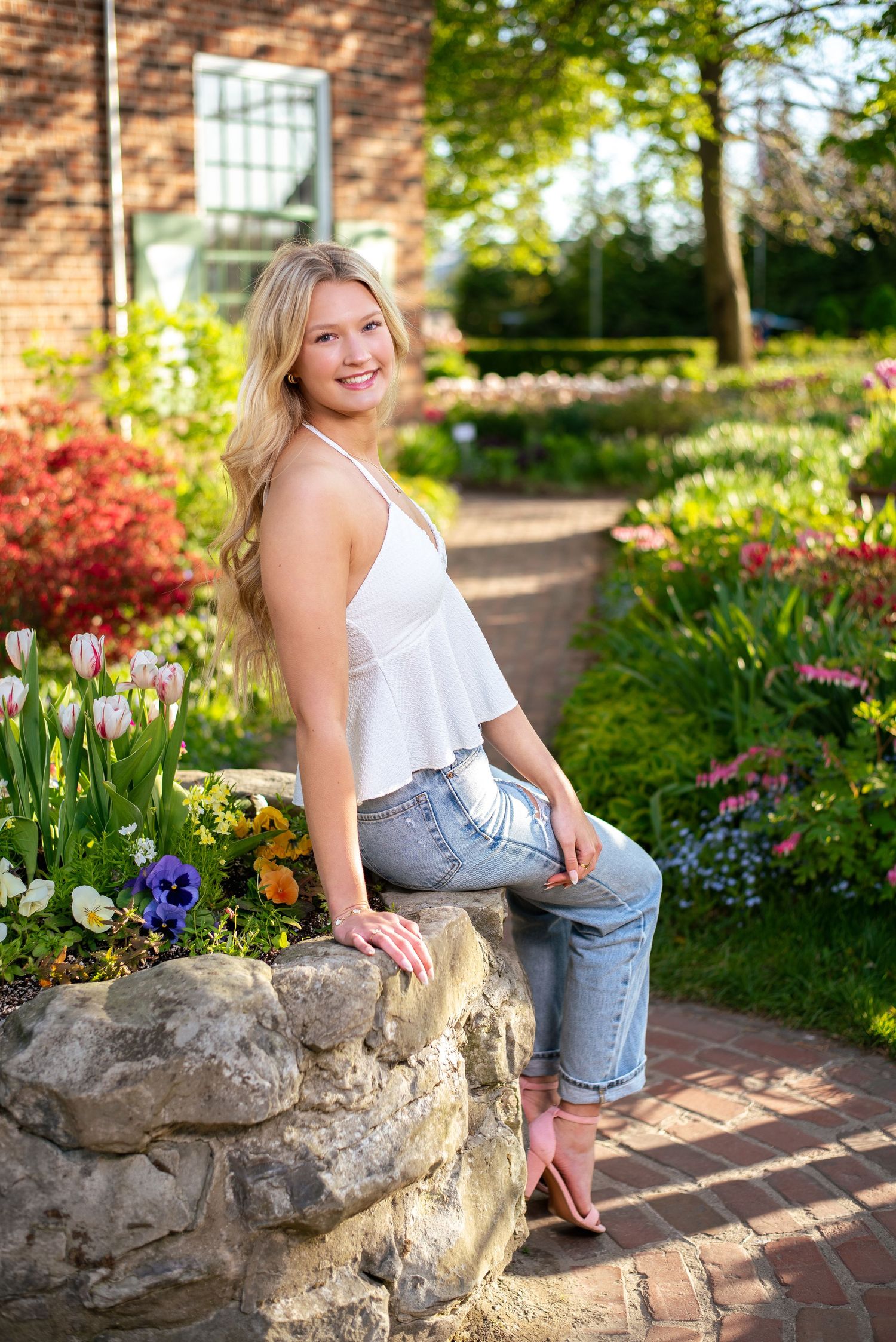 Woman in summer outfit sits on stone wall surrounded by colorful garden flowers.