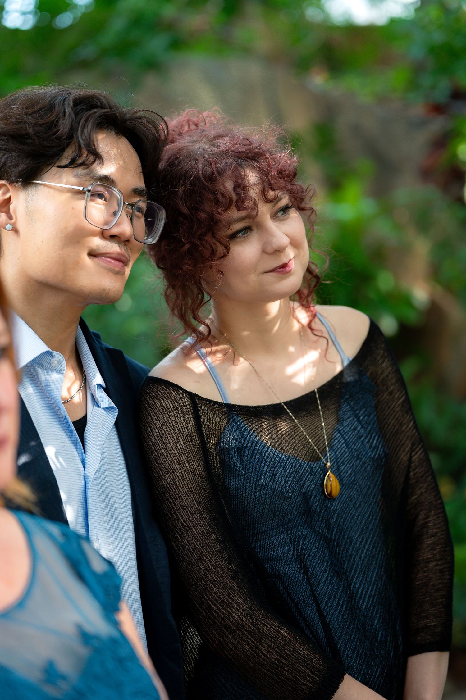 Two young people standing outdoors, one wearing glasses and a blazer, the other in a black mesh top with curly red hair.