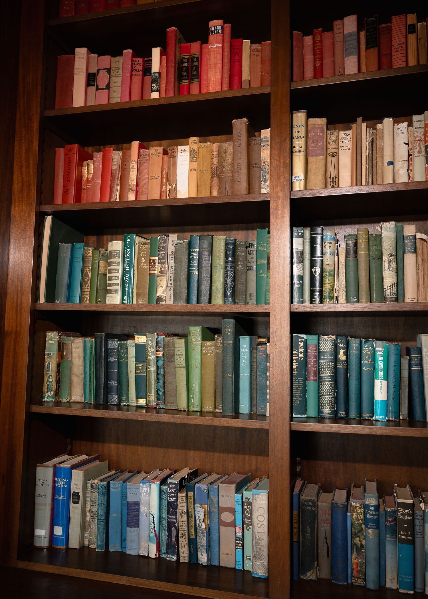 Wooden bookshelf filled with colorful vintage books arranged by color, featuring red, teal, and blue spines.