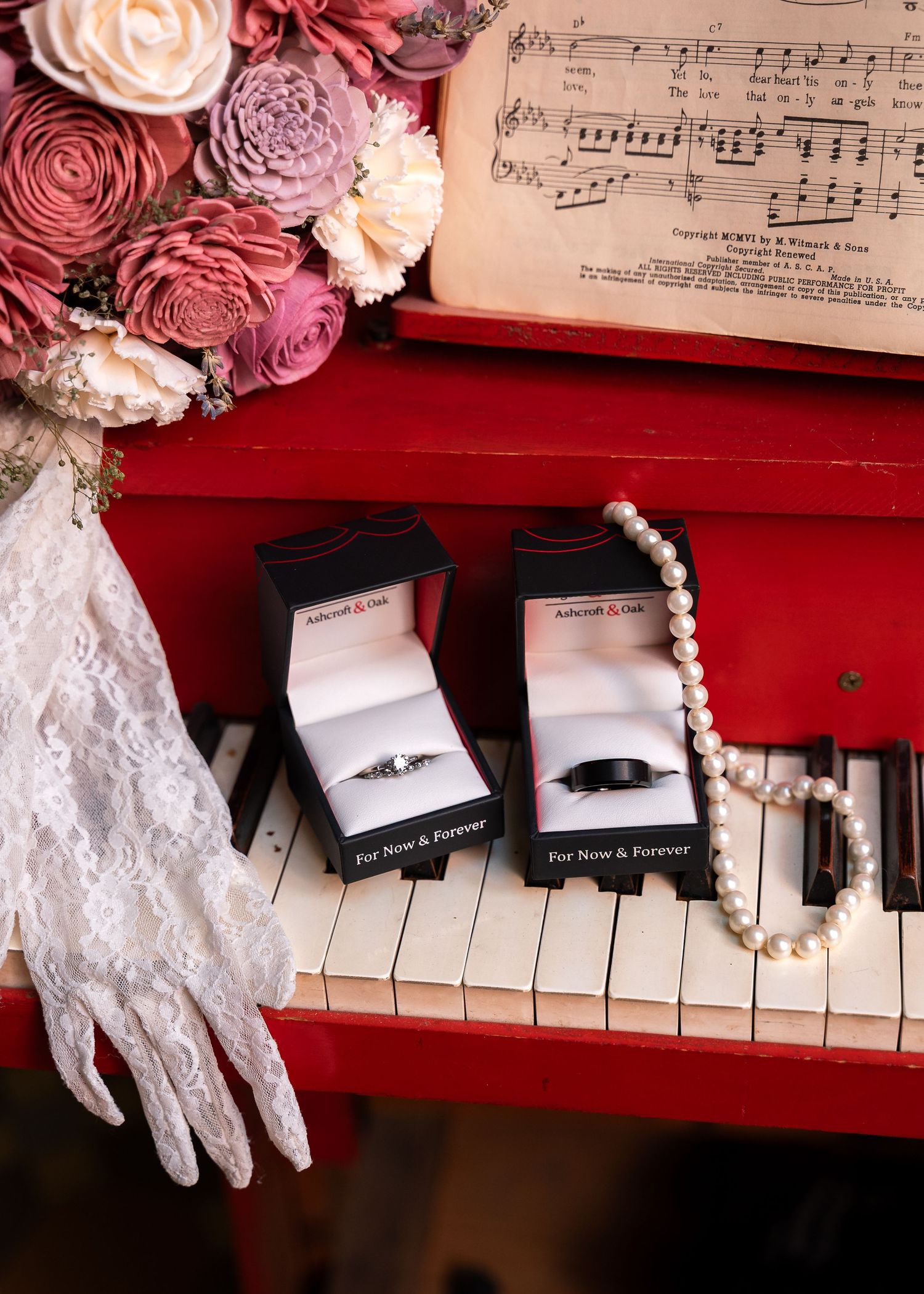 Two wedding rings in boxes on a red piano with sheet music, pearl necklace, lace glove, and pink floral bouquet.