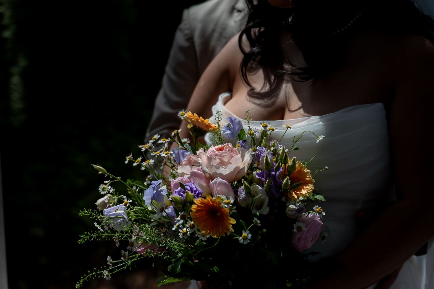 Bride holding a colorful wedding bouquet with pink roses, purple flowers, and orange gerbera daisies in a romantic outdoor setting.