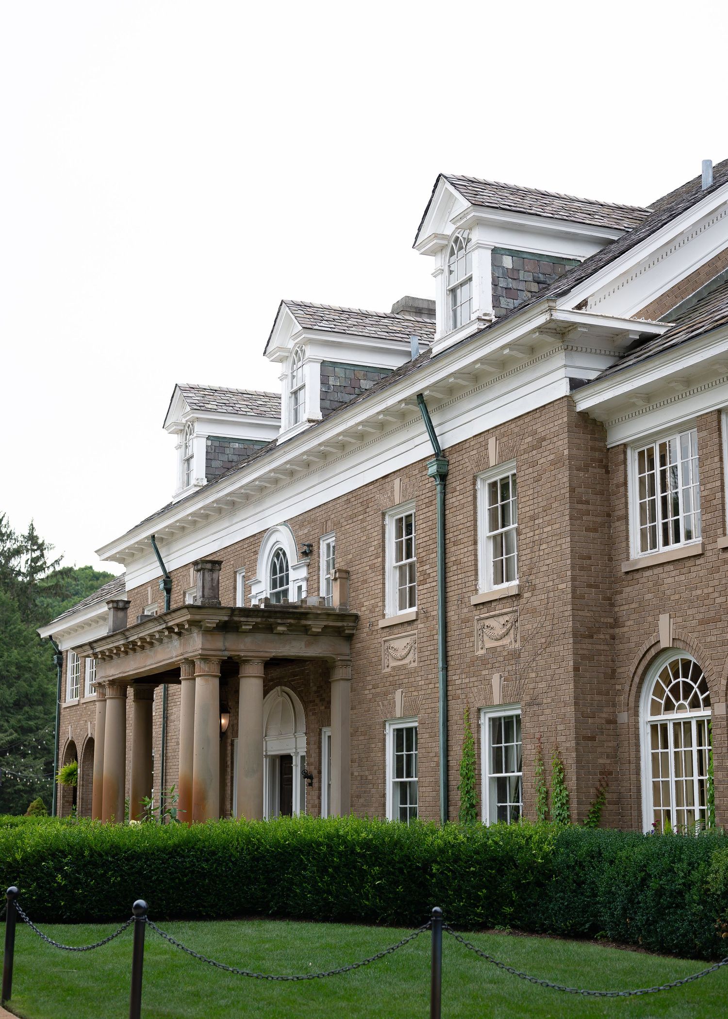 Elegant brick Georgian mansion with white trim, dormer windows, columned portico, and manicured hedges on a cloudy day.