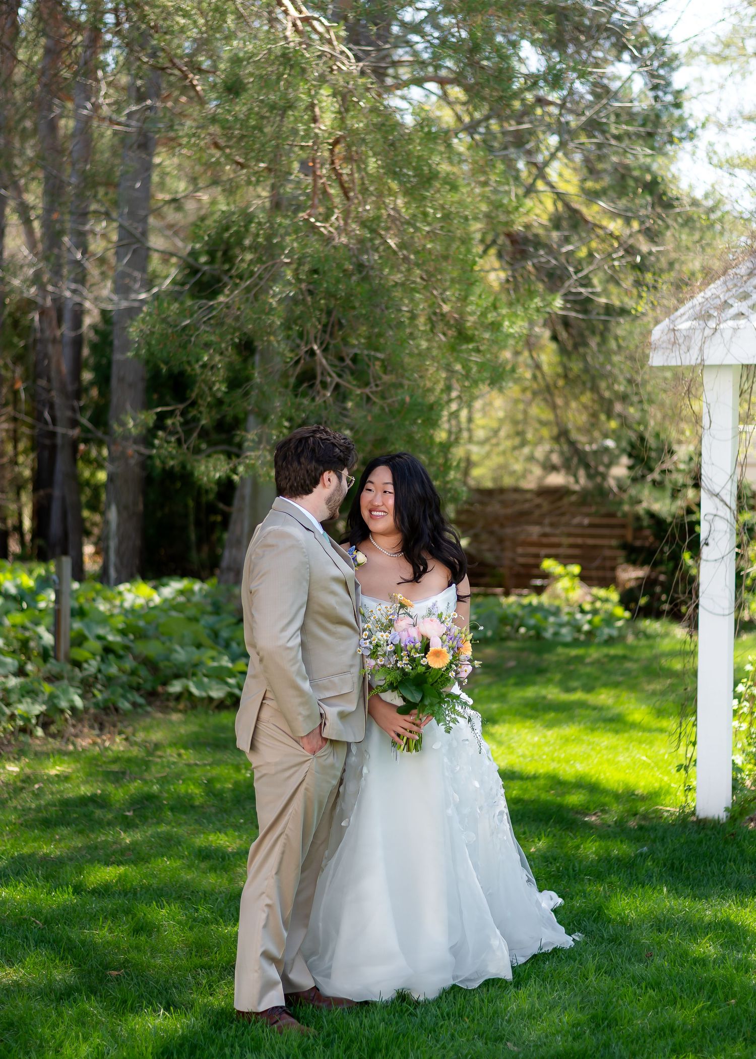 A bride and groom share a joyful moment outdoors, with the bride holding a colorful bouquet in a lush garden setting.