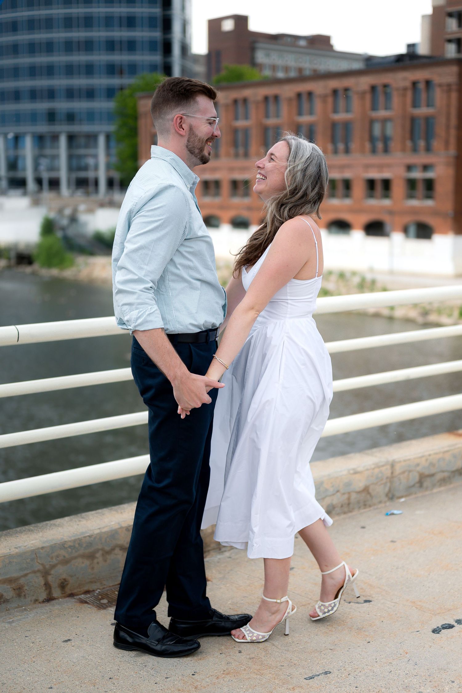 Couple holding hands and smiling on a bridge, woman in white dress and heels, man in light blue shirt and navy pants.