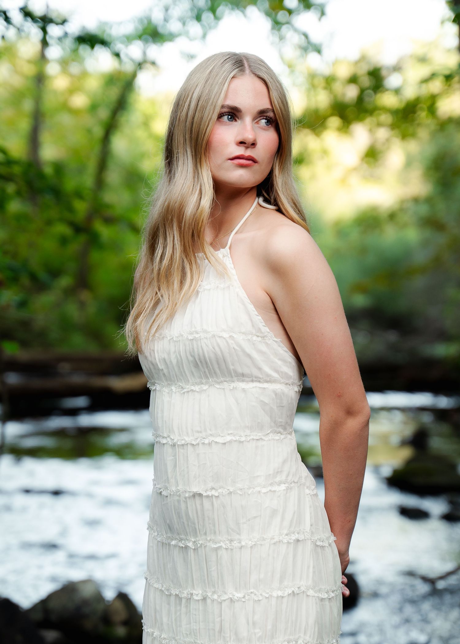Person in white tiered dress posing near flowing stream in natural setting.