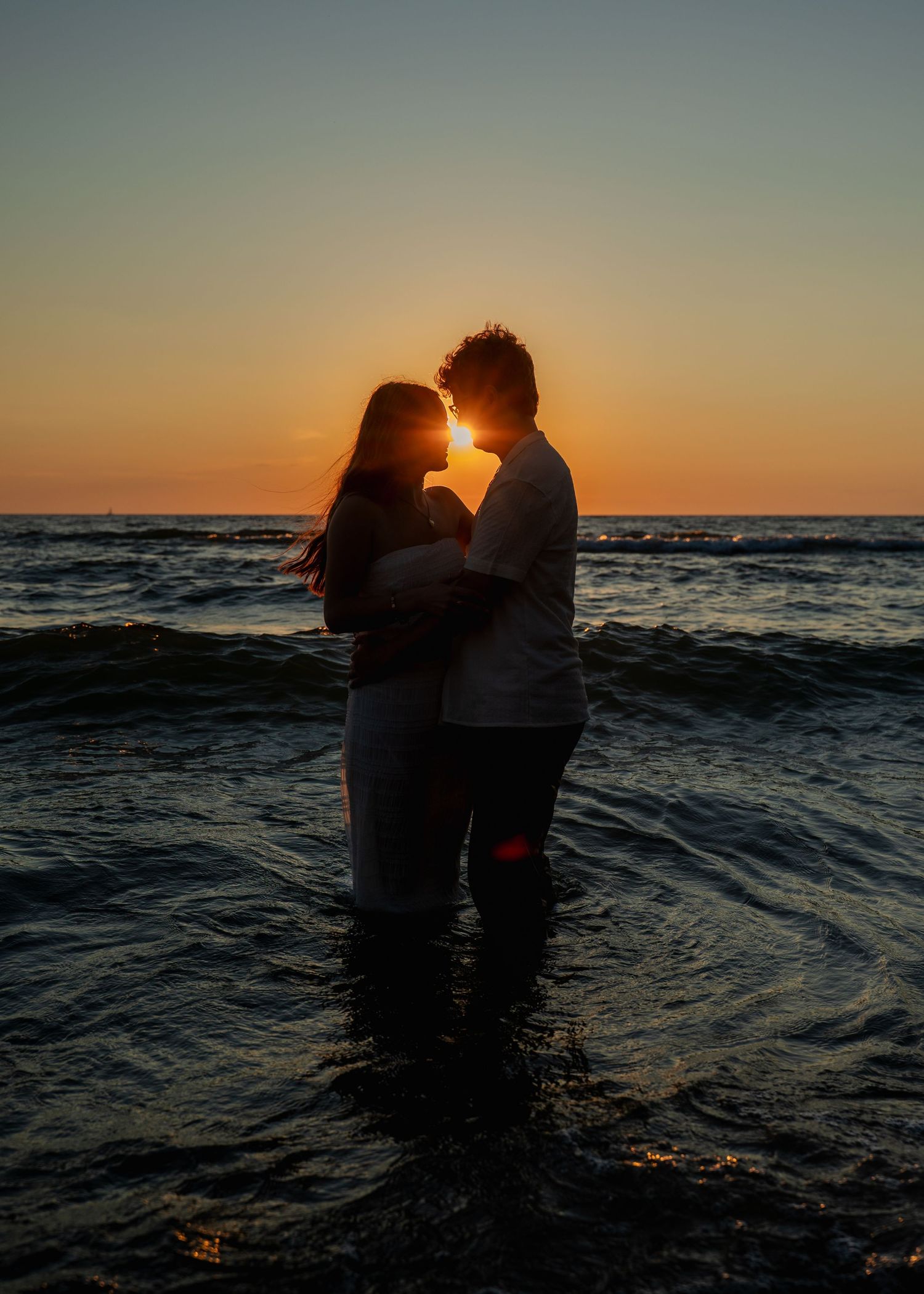 Silhouette of couple embracing in ocean water against dramatic sunset sky.