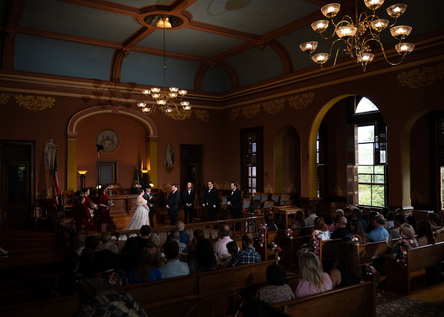 Wedding ceremony in ornate historic hall with guests seated in pews, chandelier lighting, and couple at altar.