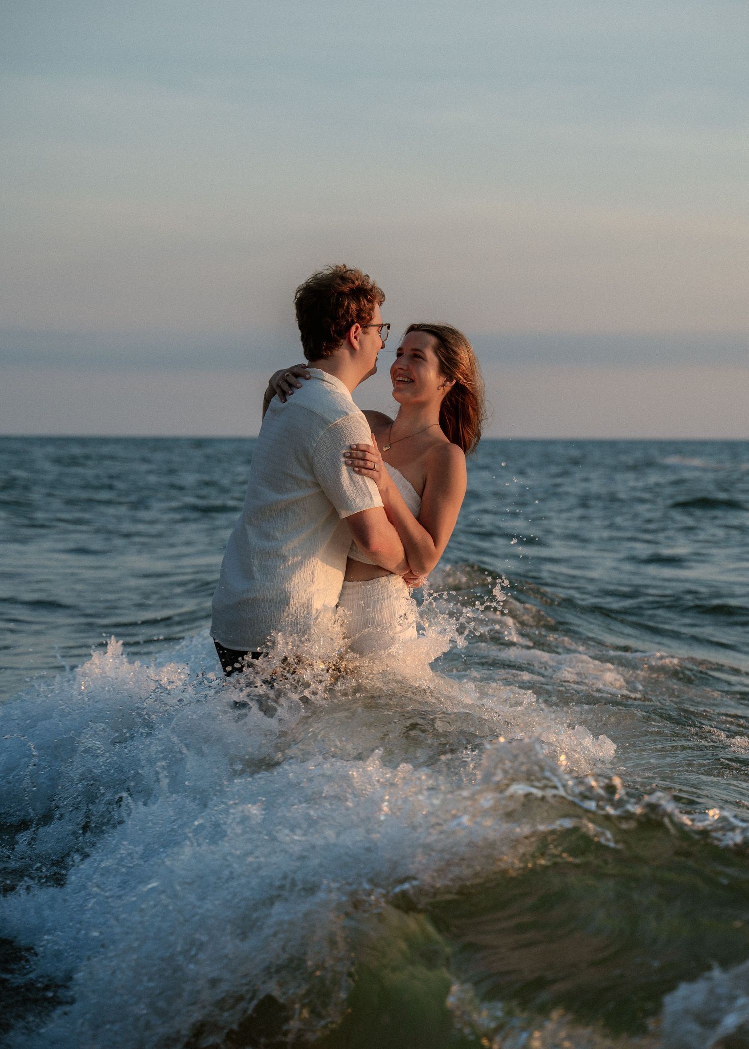 Couple embracing in ocean waves during sunset with water splashing around them.