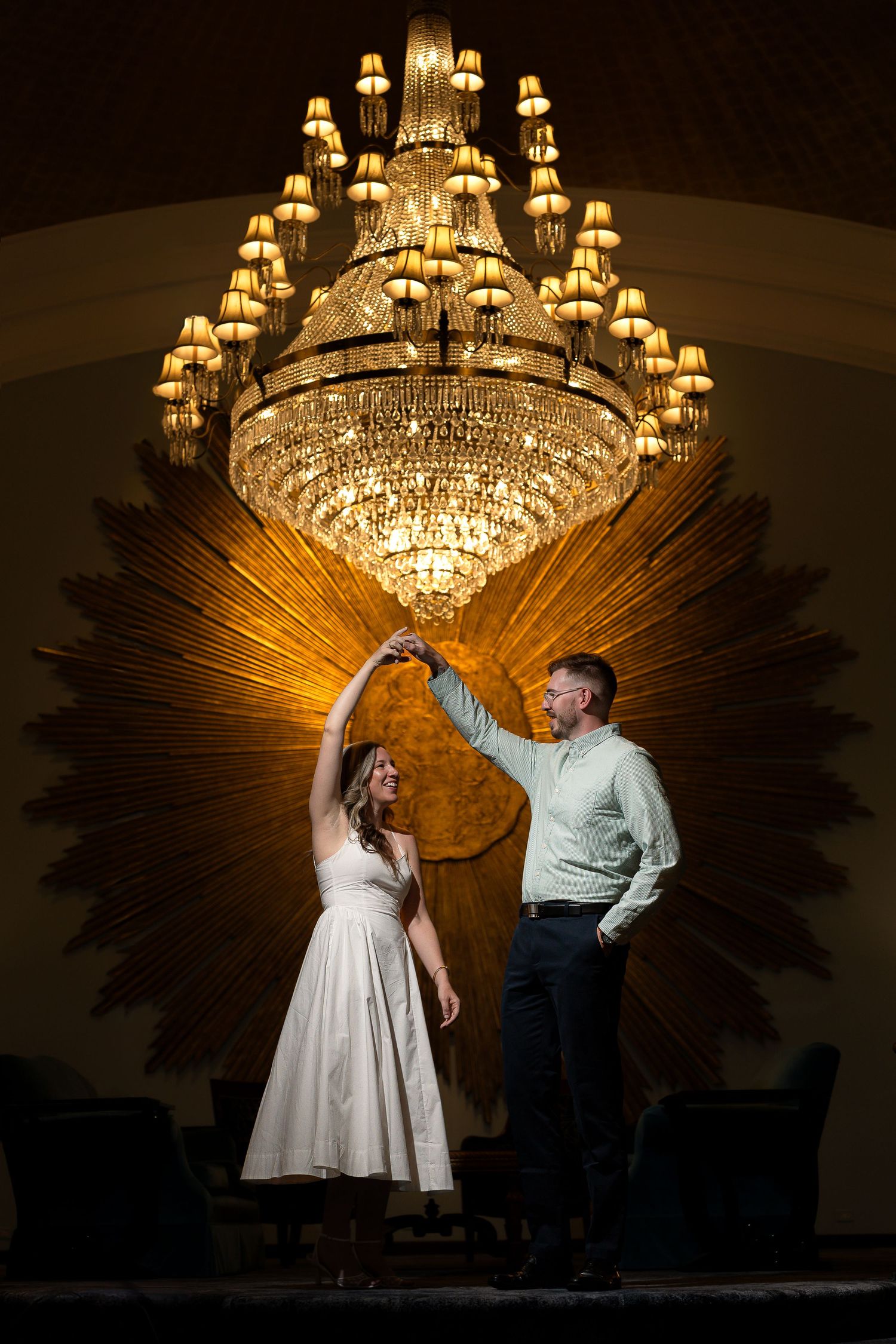 Couple dancing under elegant grand chandelier with golden sunburst decor in ornate ballroom setting.