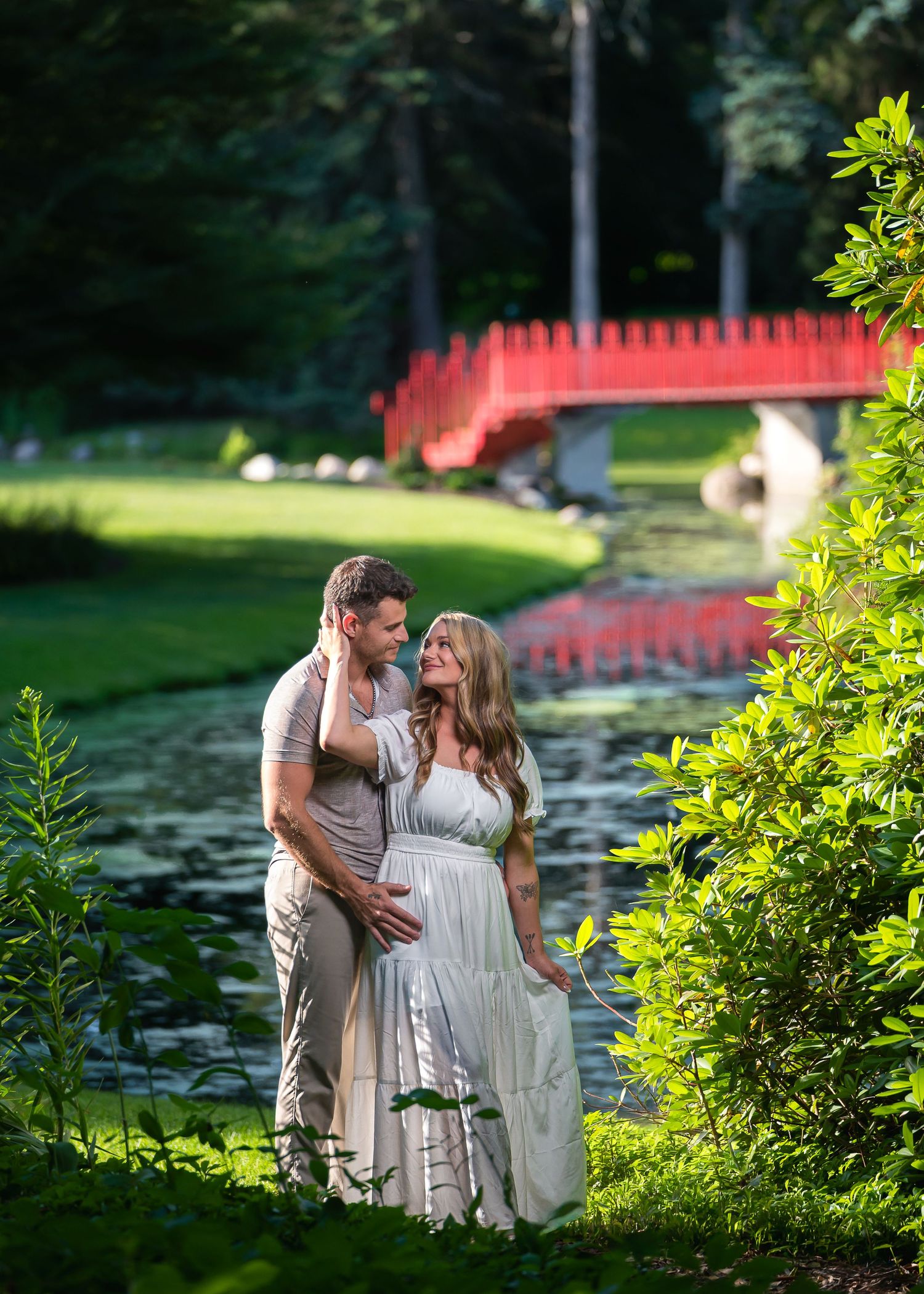 Pregnant couple embracing in lush garden with red bridge and pond backdrop.