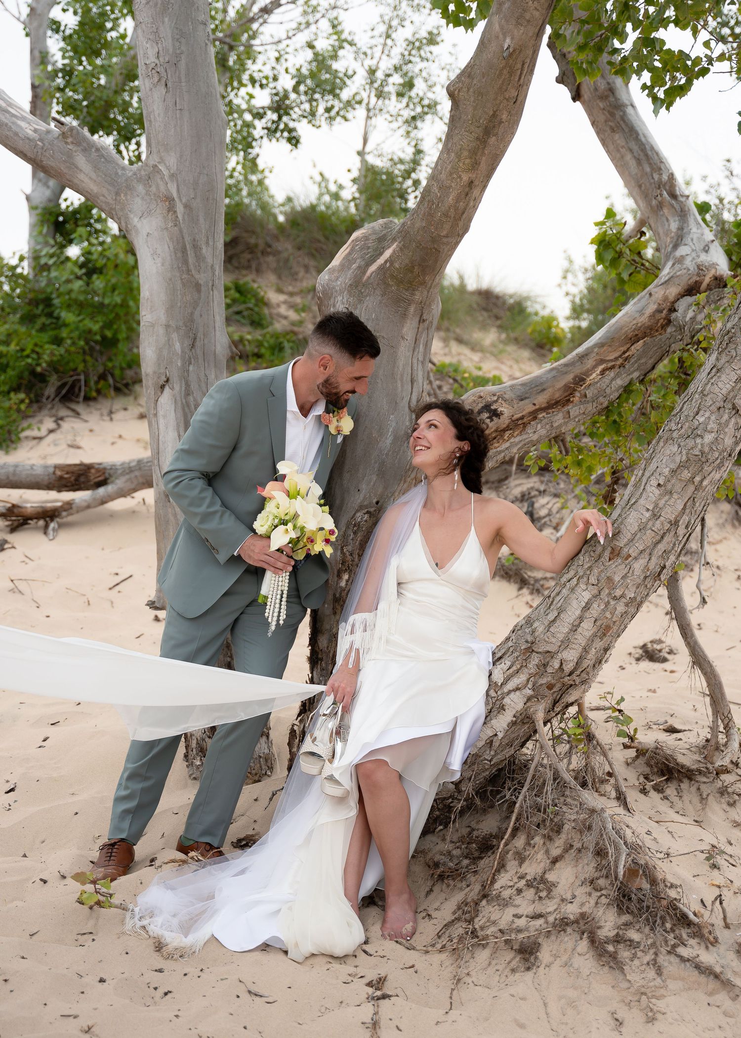 Bride and groom share a romantic moment under a driftwood tree on a sandy beach, bride holding a bouquet.