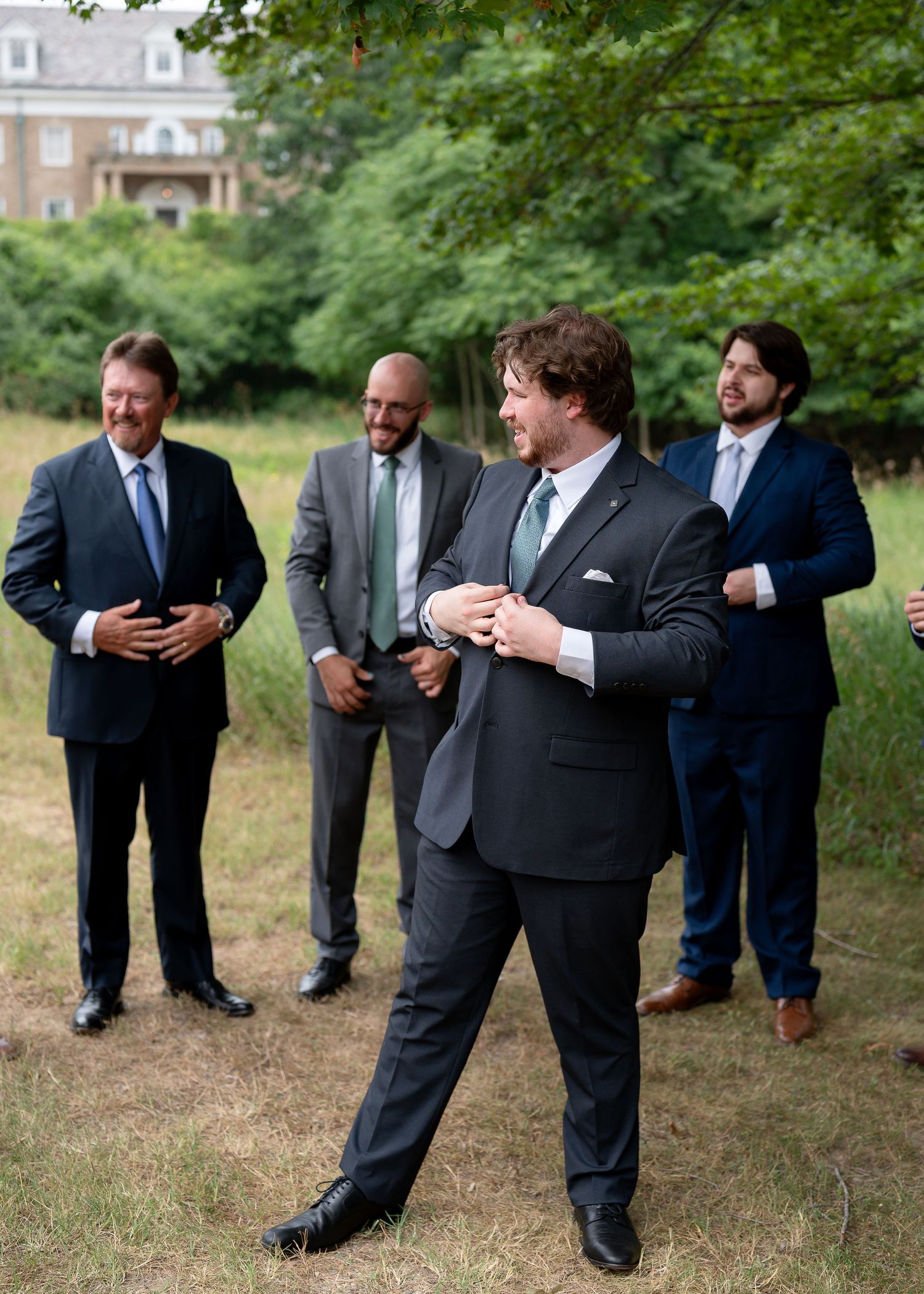 Groom adjusting suit jacket outdoors with groomsmen in navy and gray suits standing on grass.