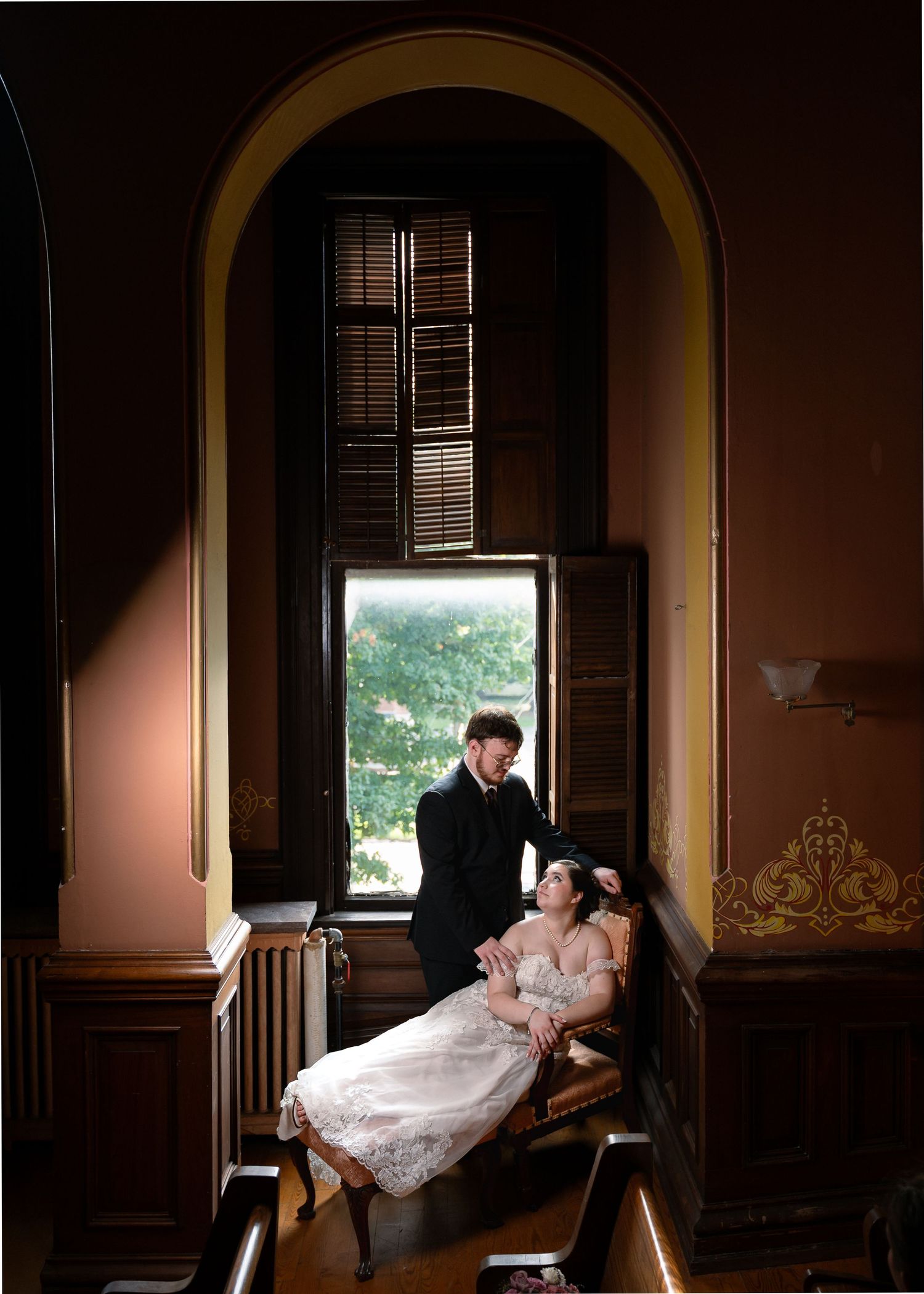 Elegant couple in wedding attire posing by an arched window in a grand Victorian-style interior.