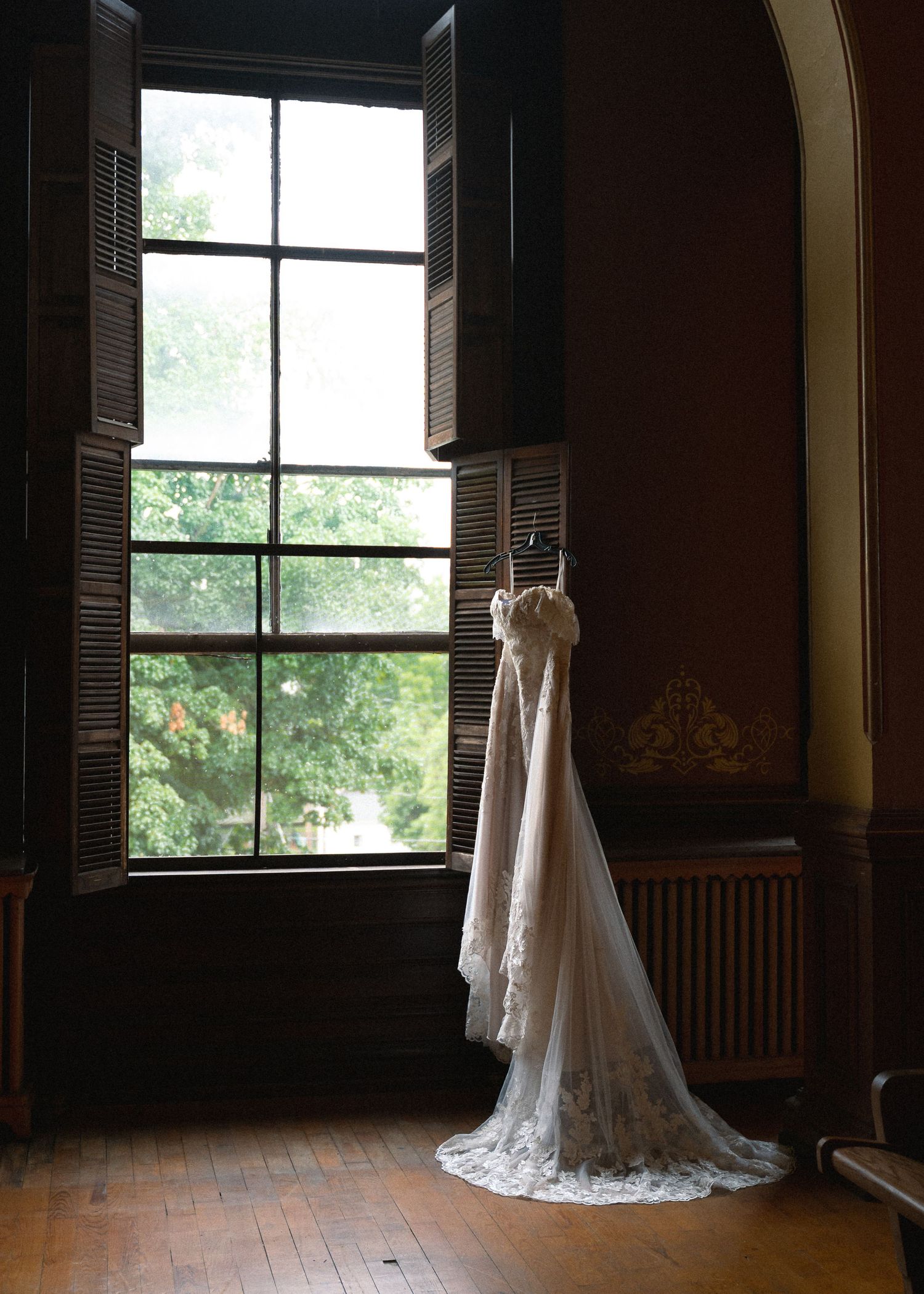 A elegant white lace wedding dress with a long train hanging by a tall window in a historic Victorian-style room.